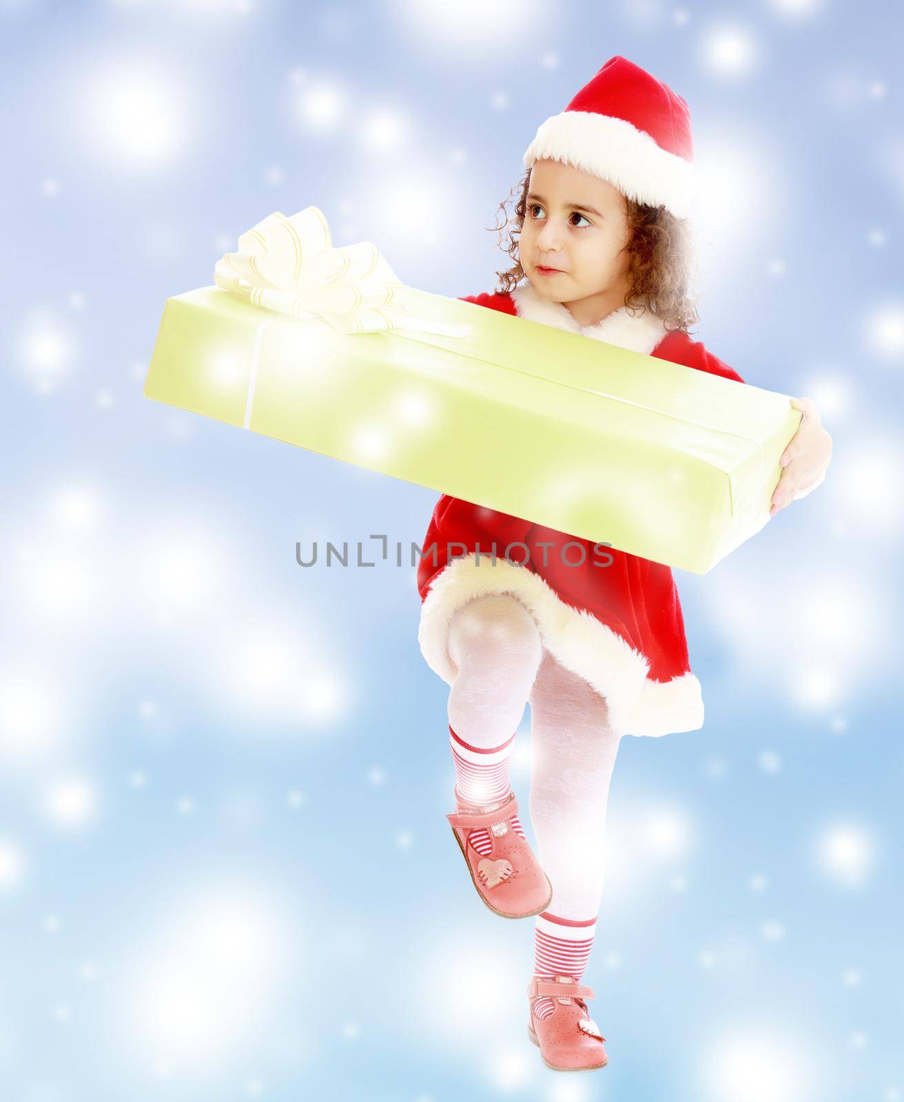 Cute little curly girl in a coat and hat of Santa Claus carries the big green box. A girl holds a box with his knee.Blue winter background with white snowflakes.