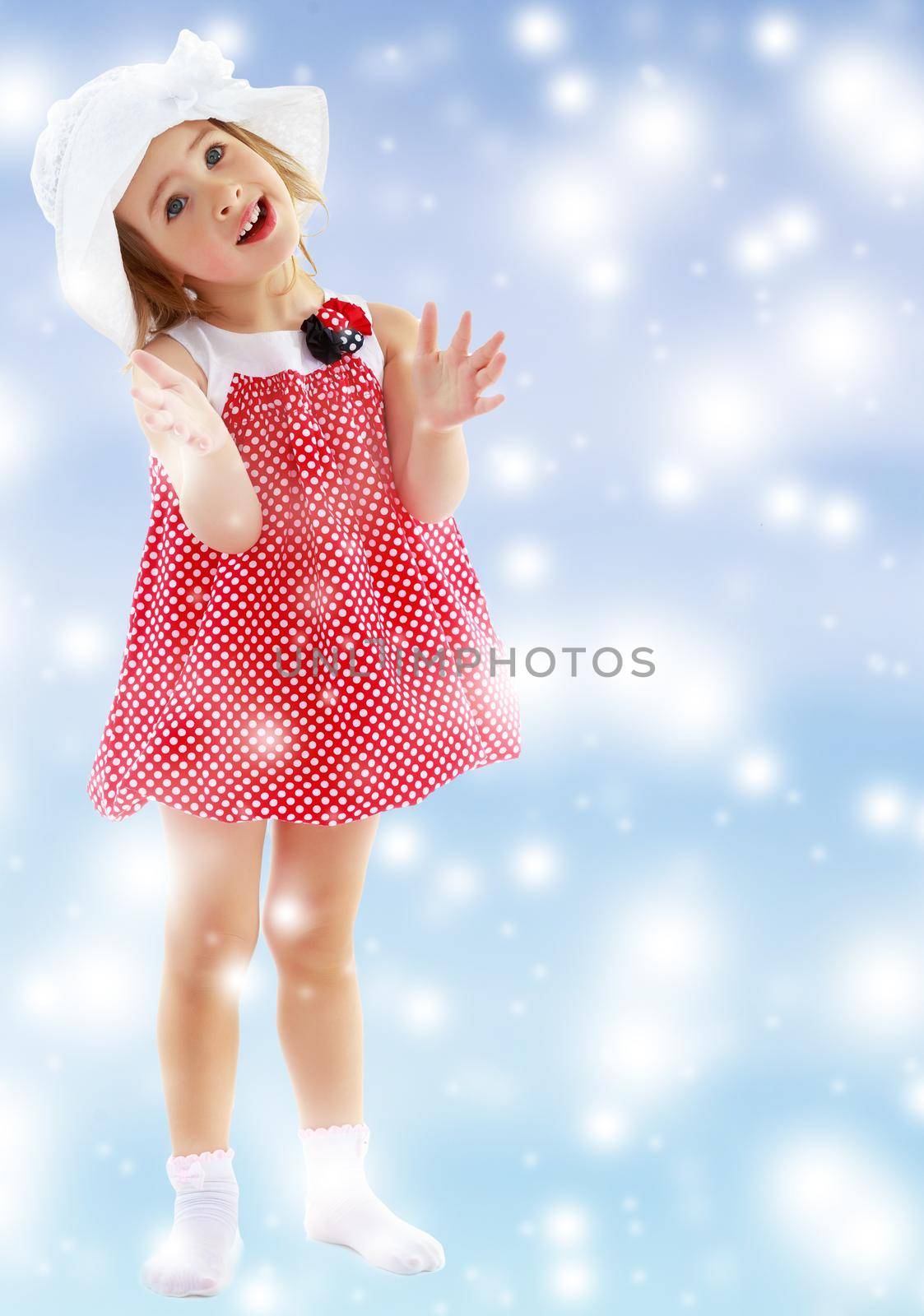Joyful little girl in a very short polka dot dress and white Panama city beach , claps.Full growth.Gentle blue Christmas background with white snowflakes abstract.