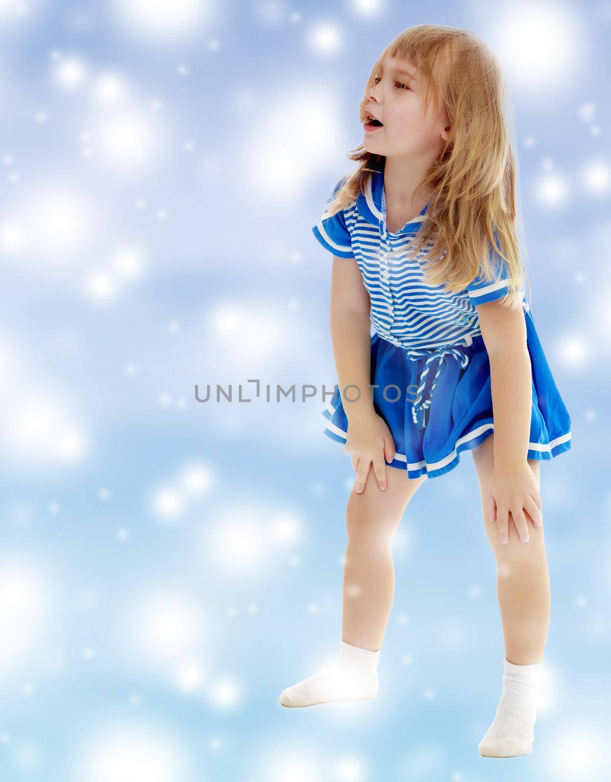 On a blue background with white blurry circles, like Christmas snowflakes. Cute little unkempt girl in a short blue dress. Girl looking to the side with his hands on his knees.