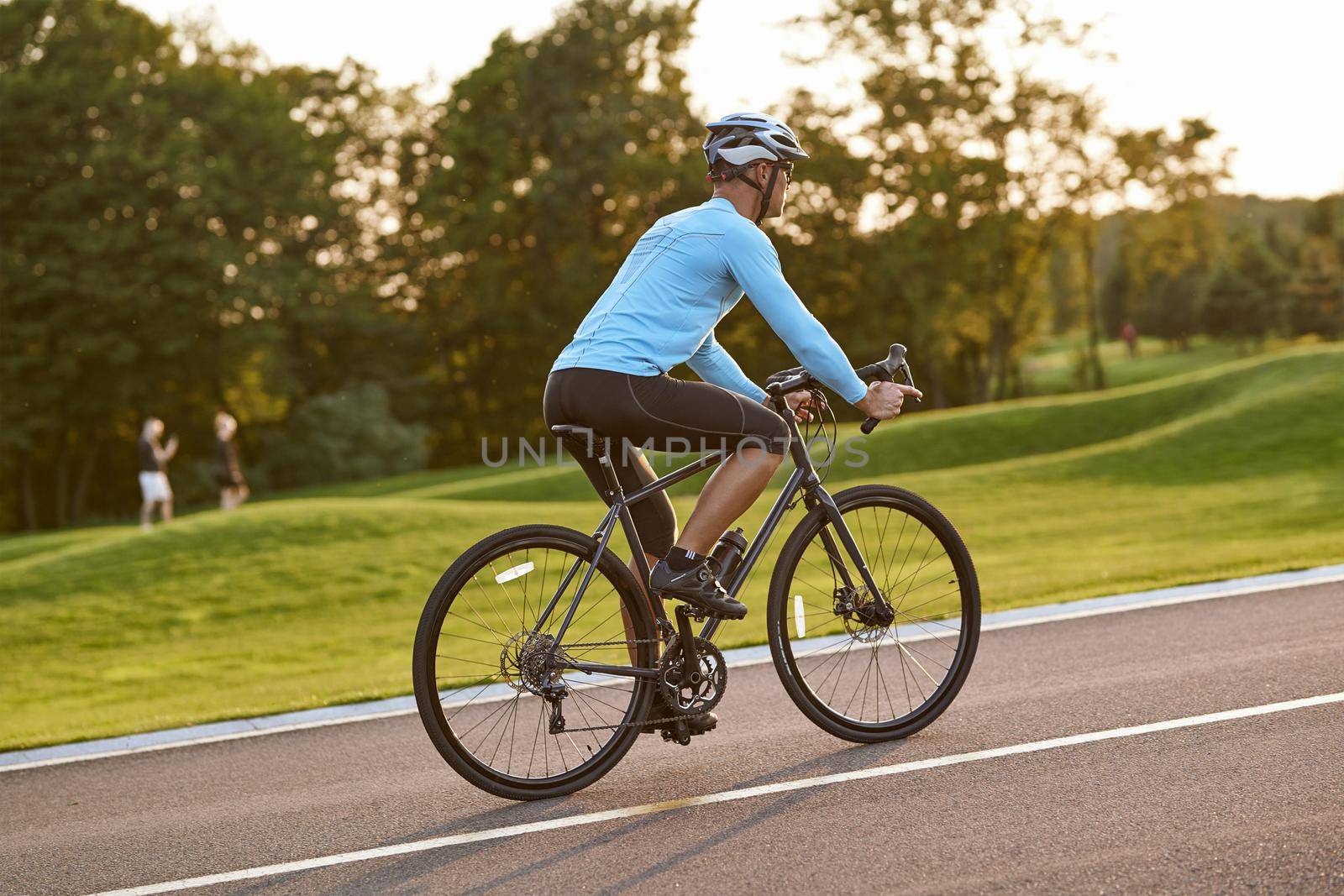 Side view of athletic man in sportswear riding bicycle along a road in city park at sunset by friendsstock