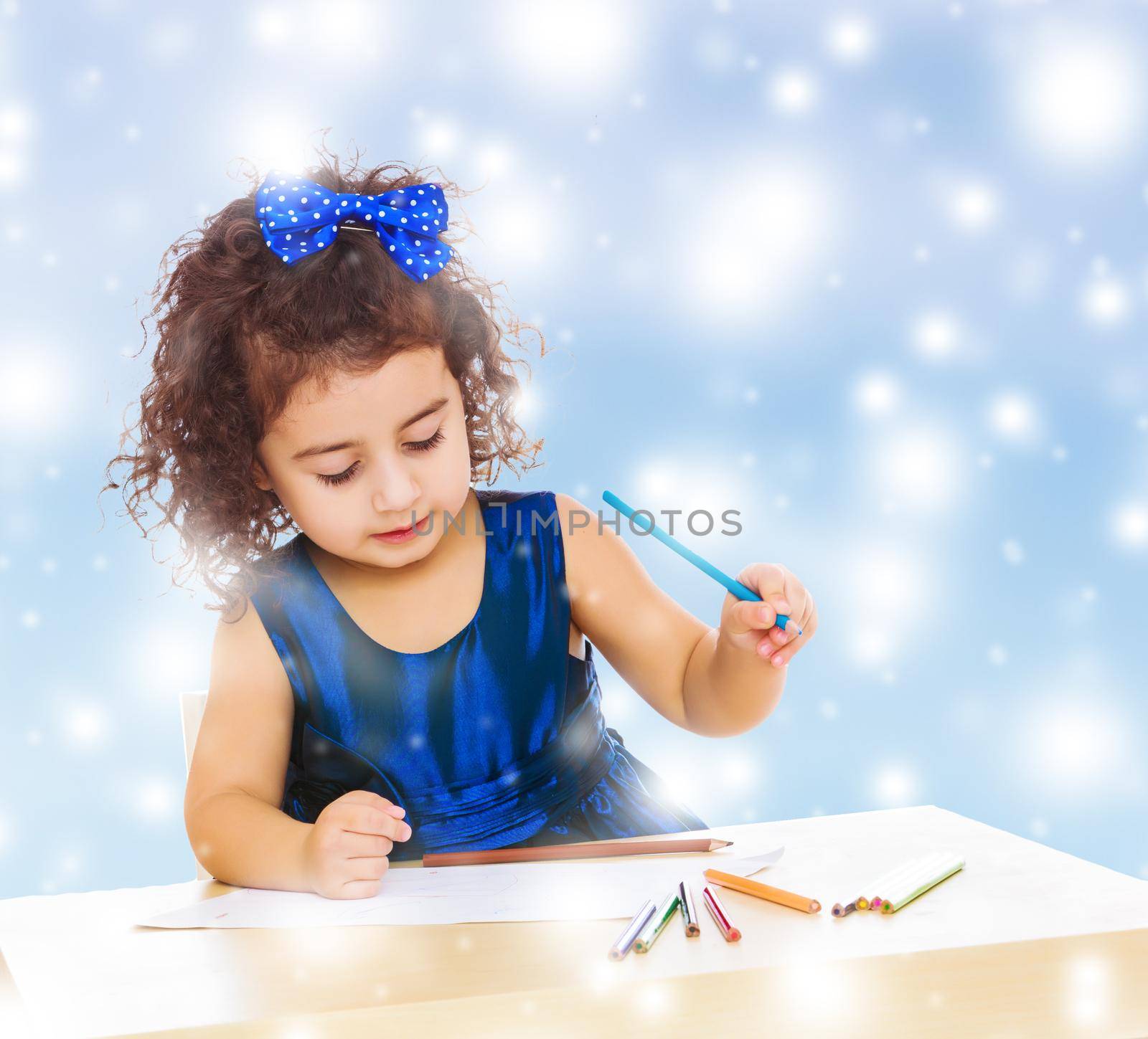 Pensive little girl in a blue dress, holding a pencil . She paints at a table in a Montessori kindergarten.Blue winter background with white snowflakes.