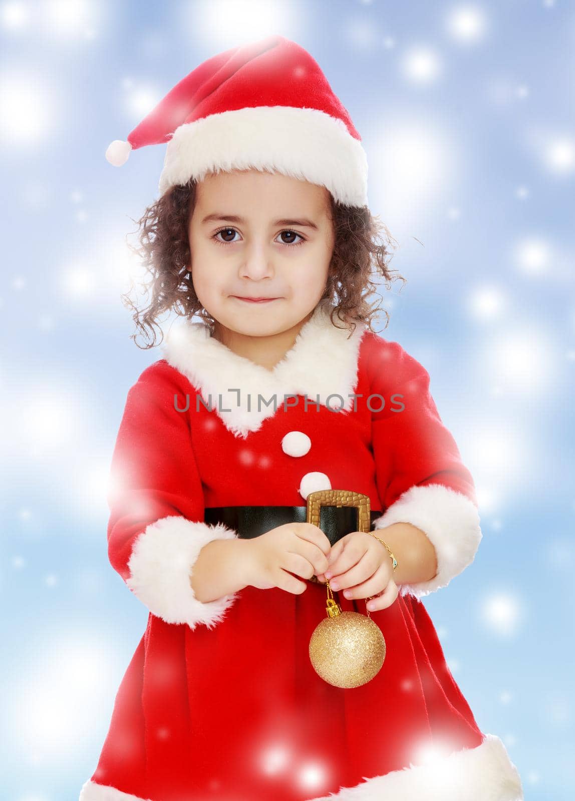 Very cute ,with curly hair little girl in a suit and cap of Santa Claus , is holding a Christmas toy. Close-up.Blue winter background with white snowflakes.
