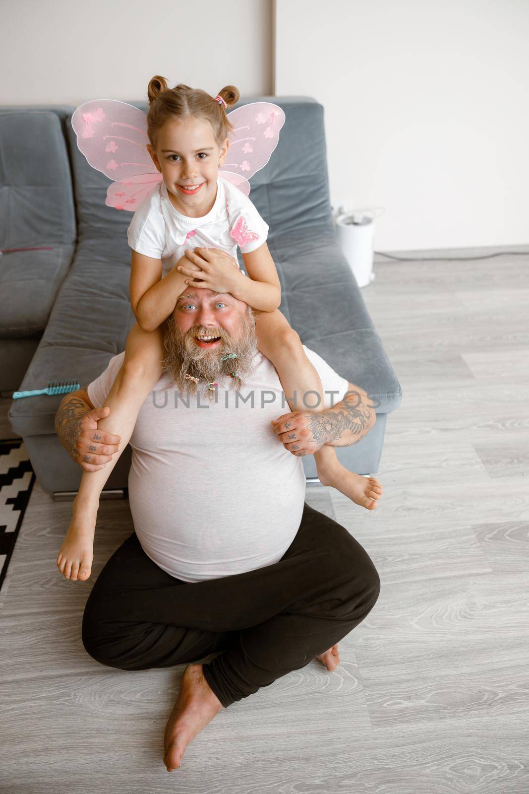Joyful little girl with pink fairy wings sits on shoulders of funny plump father with scrunchies in beard near sofa in living room. Family playtime