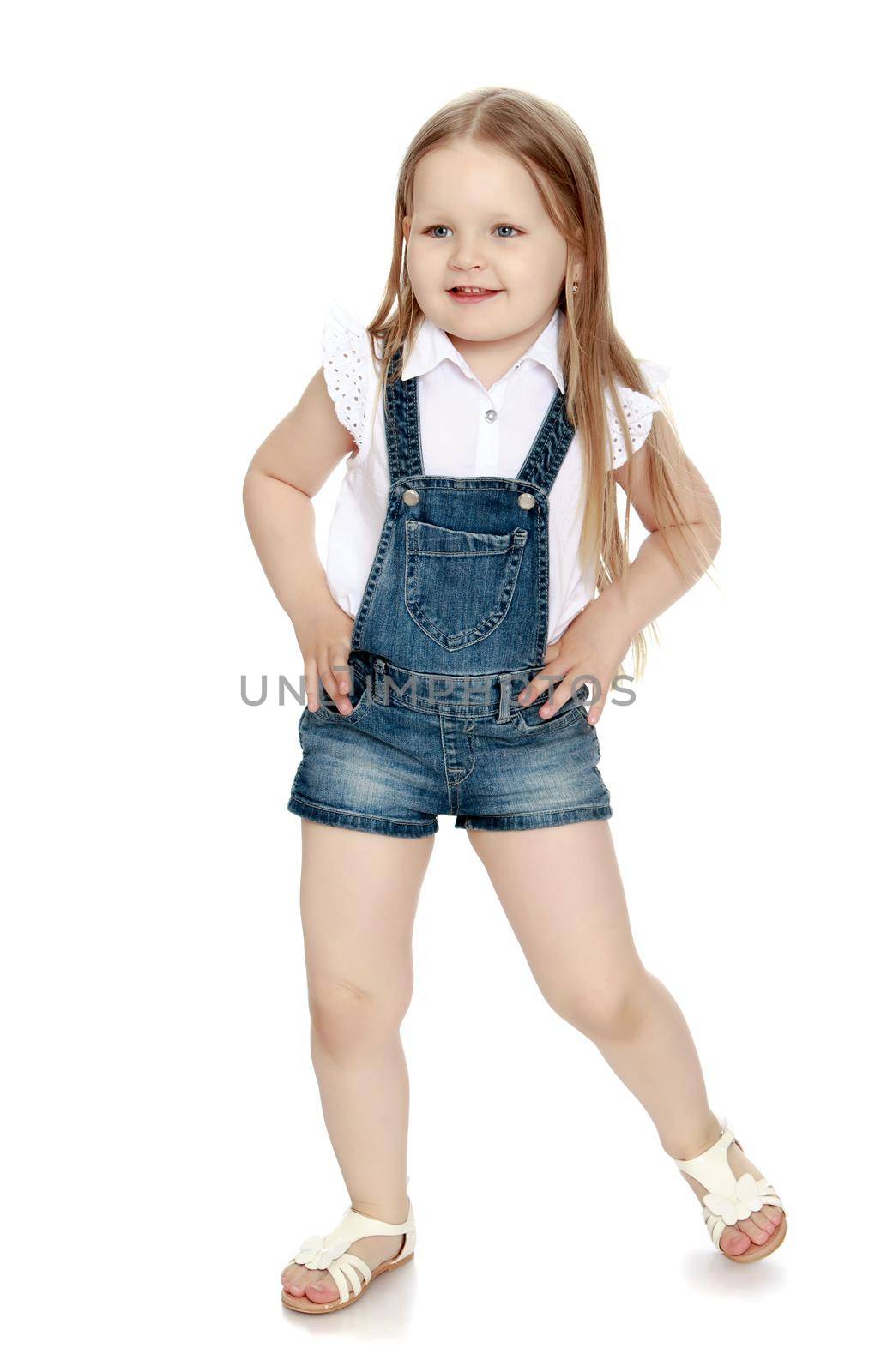 Beautiful little girl with long blonde hair below the belt in short denim jumpsuit. Girl holding hands on waist - Isolated on white background