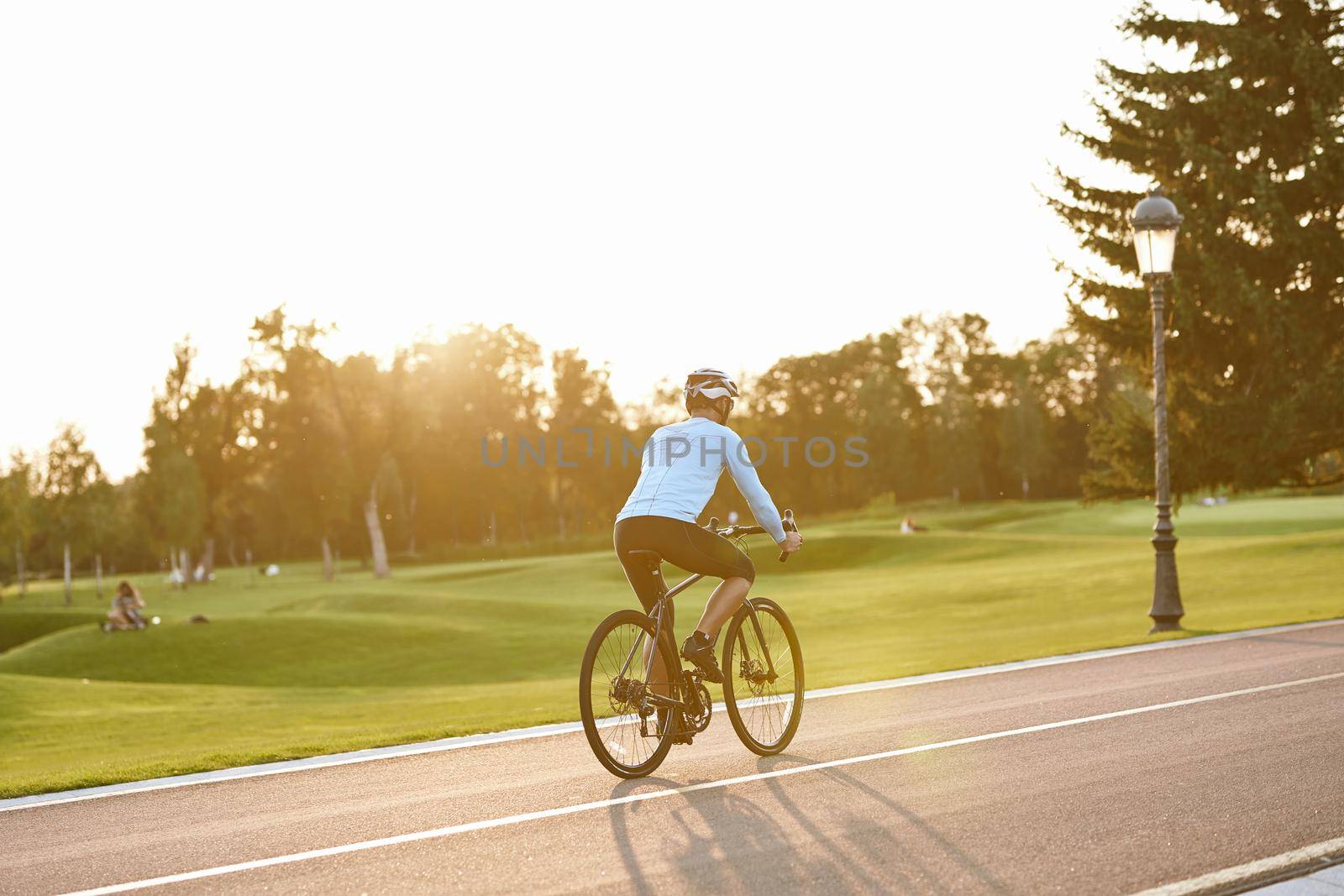Perfect day for cycling. Rear view of athletic man in sportswear riding bicycle along the road in city park at sunset, full length. Healthy active lifestyle and sport concept