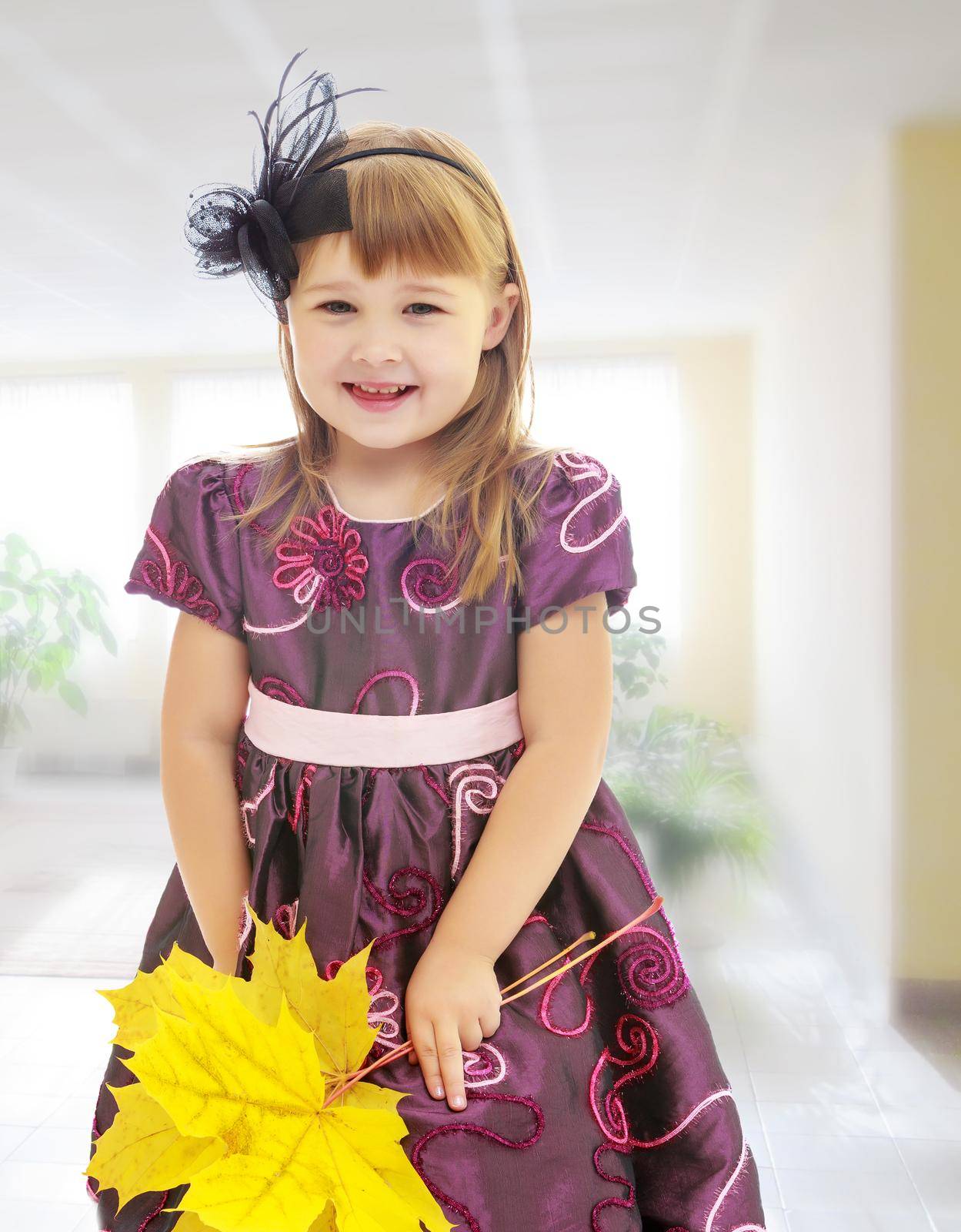 The concept of child rearing in the family and kindergarten. On the background of the hall with large , bright Windows. Nice little girl dressed in brown dress. She is holding a bouquet of maple leaves.