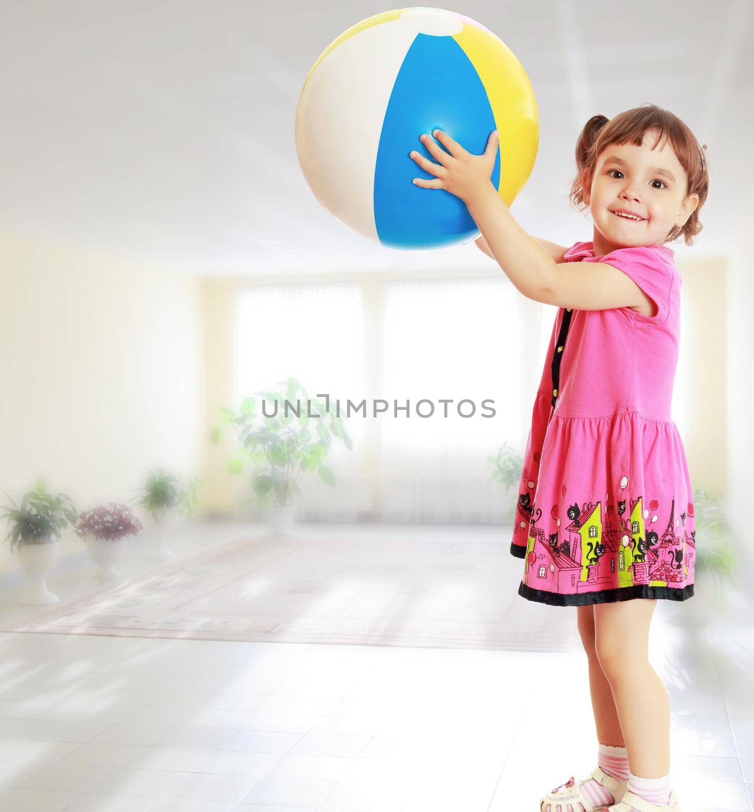 Happy little girl with pigtails on the head , in a pink dress. The girl lifted a large, inflatable striped ball.Against the background of a large room with Windows and flowers .