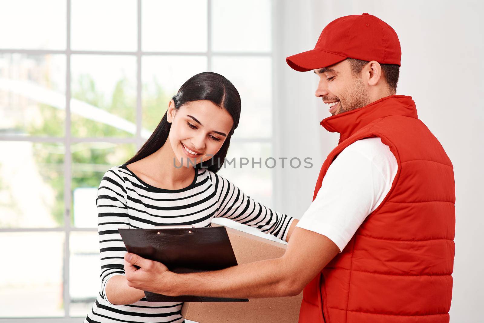 Cheerful man wearing red postal uniform is delivering parcel to a satisfied client. Dark-haired woman is signing receipt of delivery package. Friendly worker, high quality delivery service. Indoors.