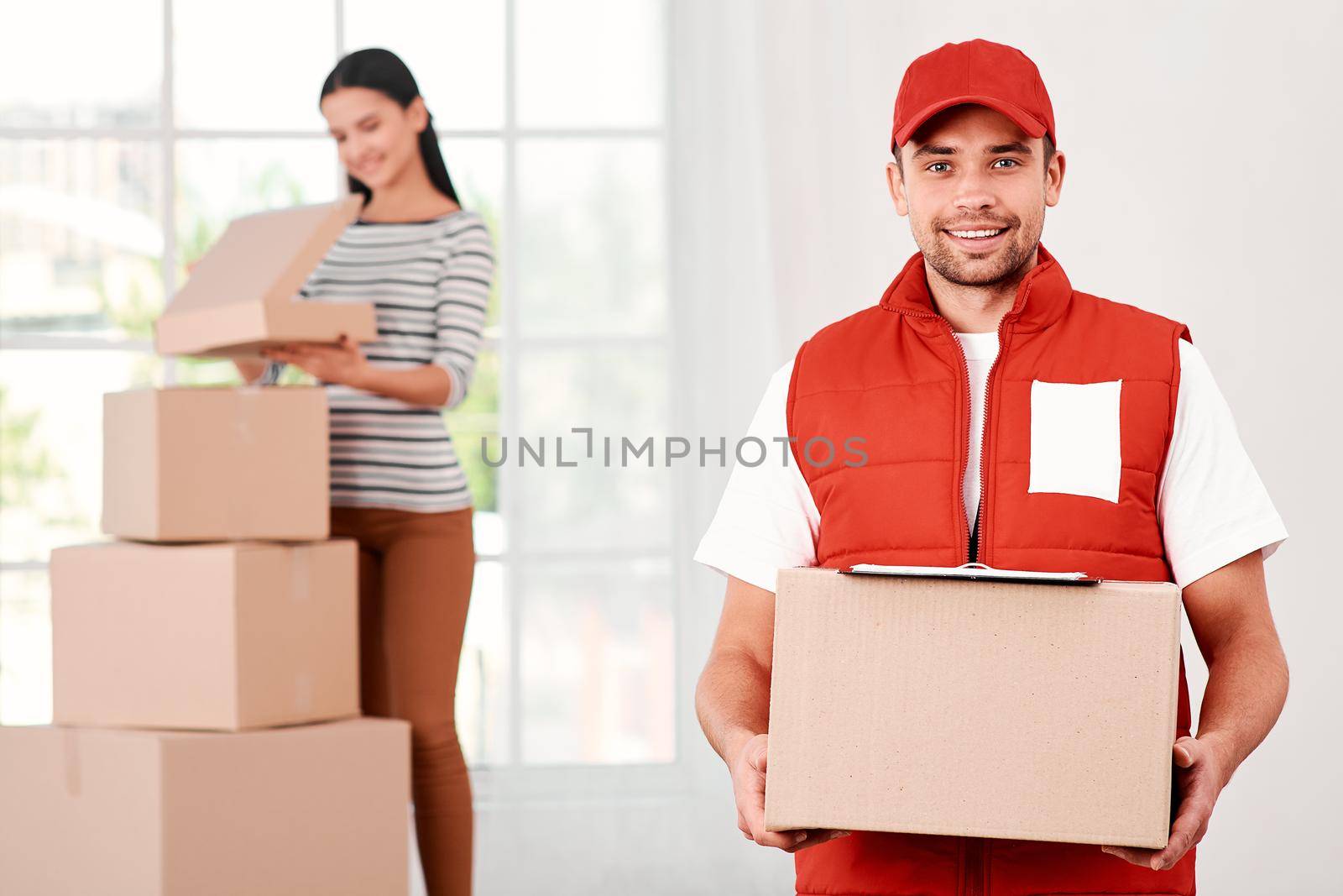 Young courier in red postal uniform standing with parcels indoors, looking at the camera with a smile. Woman unpacking her purchases in the background. A stack of carton boxes. Bright interior. Friendly worker, high quality delivery service