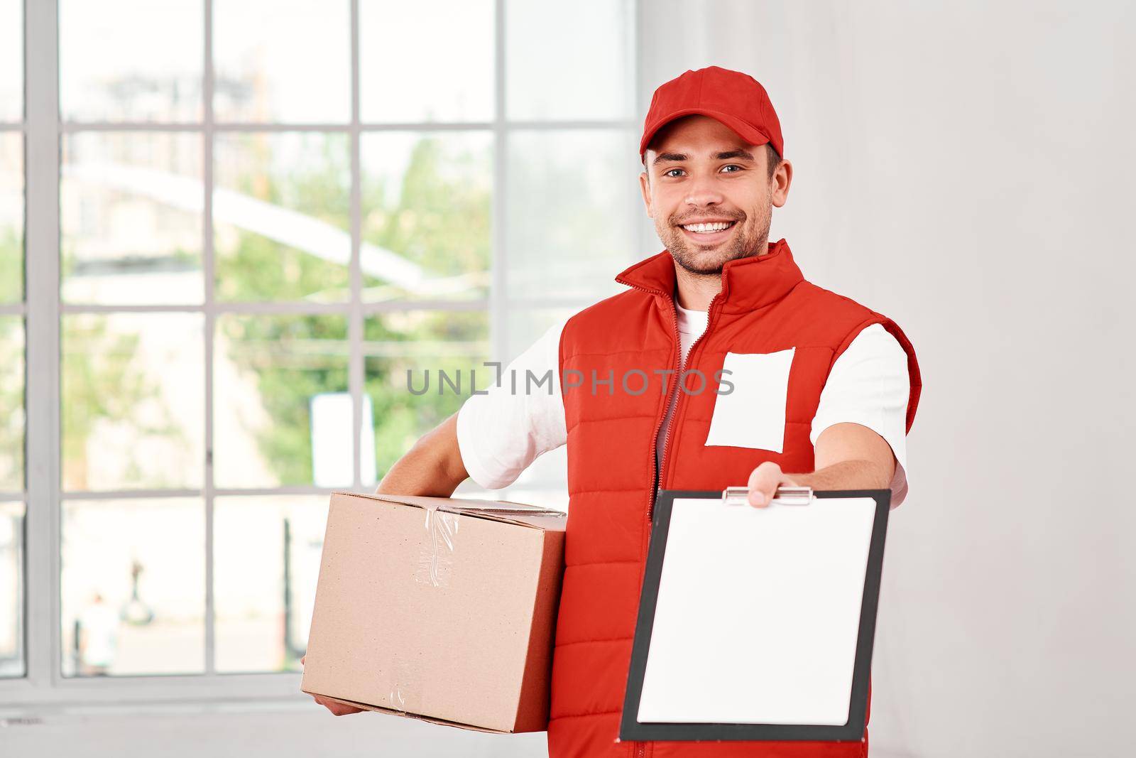 Cheerful postman wearing red postal uniform is delivering parcel to a client. He holds carton box, waiting for a signature on delivery and looking at the camera with a smile. Friendly worker, high quality delivery service. Indoors. Bright interior.