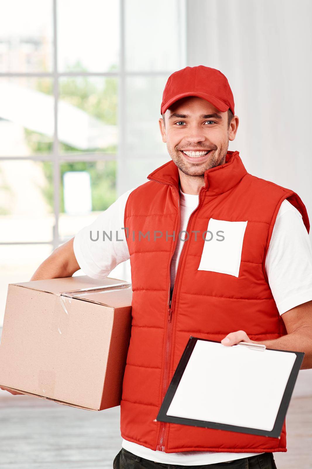 Cheerful postman wearing red postal uniform is delivering parcel to a client. He holds carton box, waiting for a signature on delivery and looking at the camera with a smile. Friendly worker, high quality delivery service. Indoors. Bright interior.