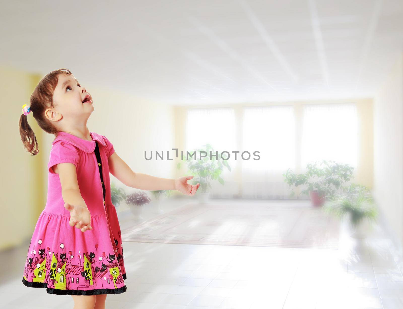 Adorable little girl with pigtails on the head , in a pink dress. The girl was looking at the top turned sideways to the camera.Against the background of a large room with Windows and flowers .