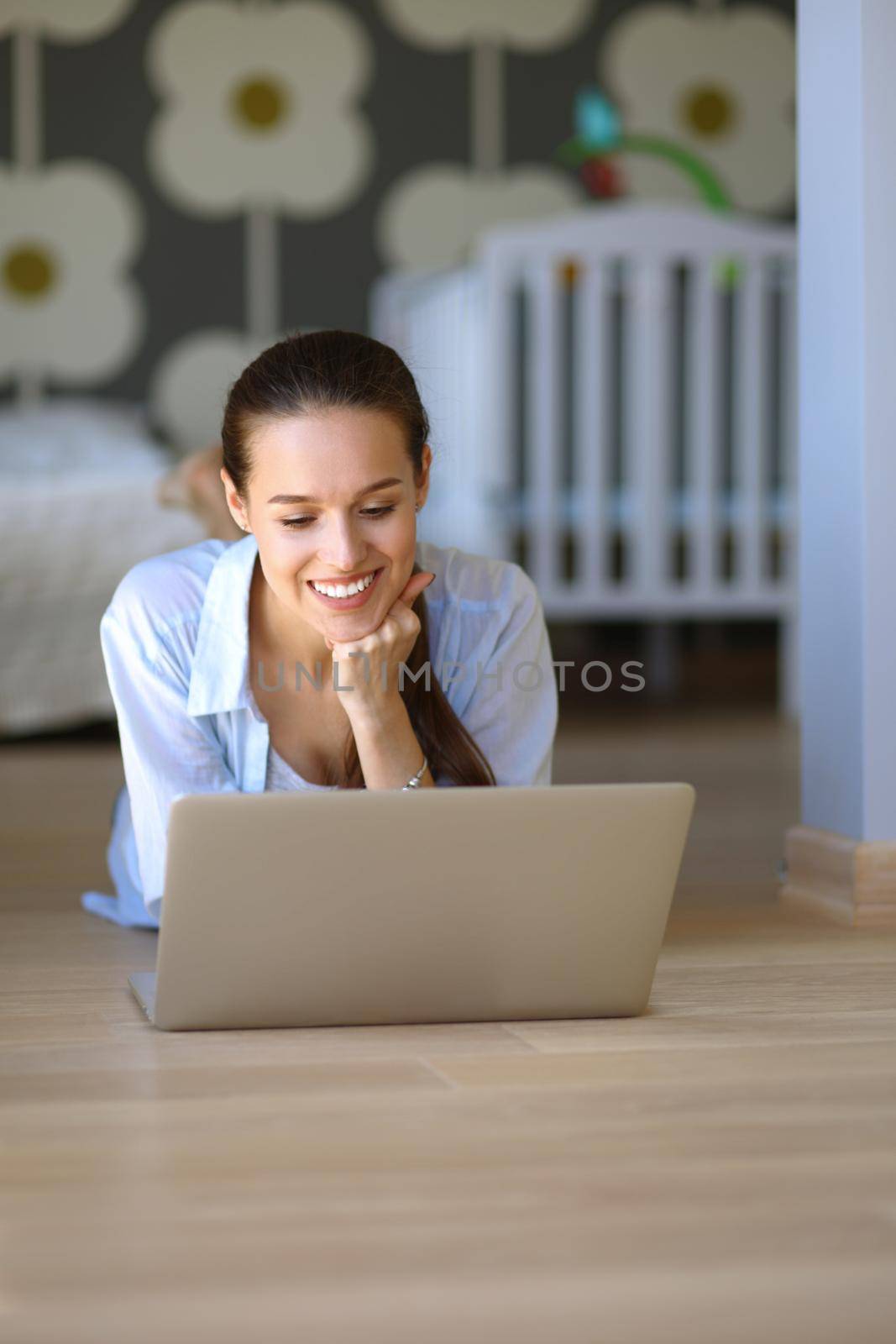 Young woman sitting on the floor near children's cot with laptop. Young mom.