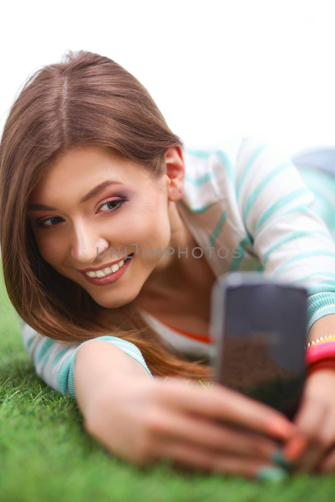 Beautiful young woman making selfie by her phone while lying in green grass.