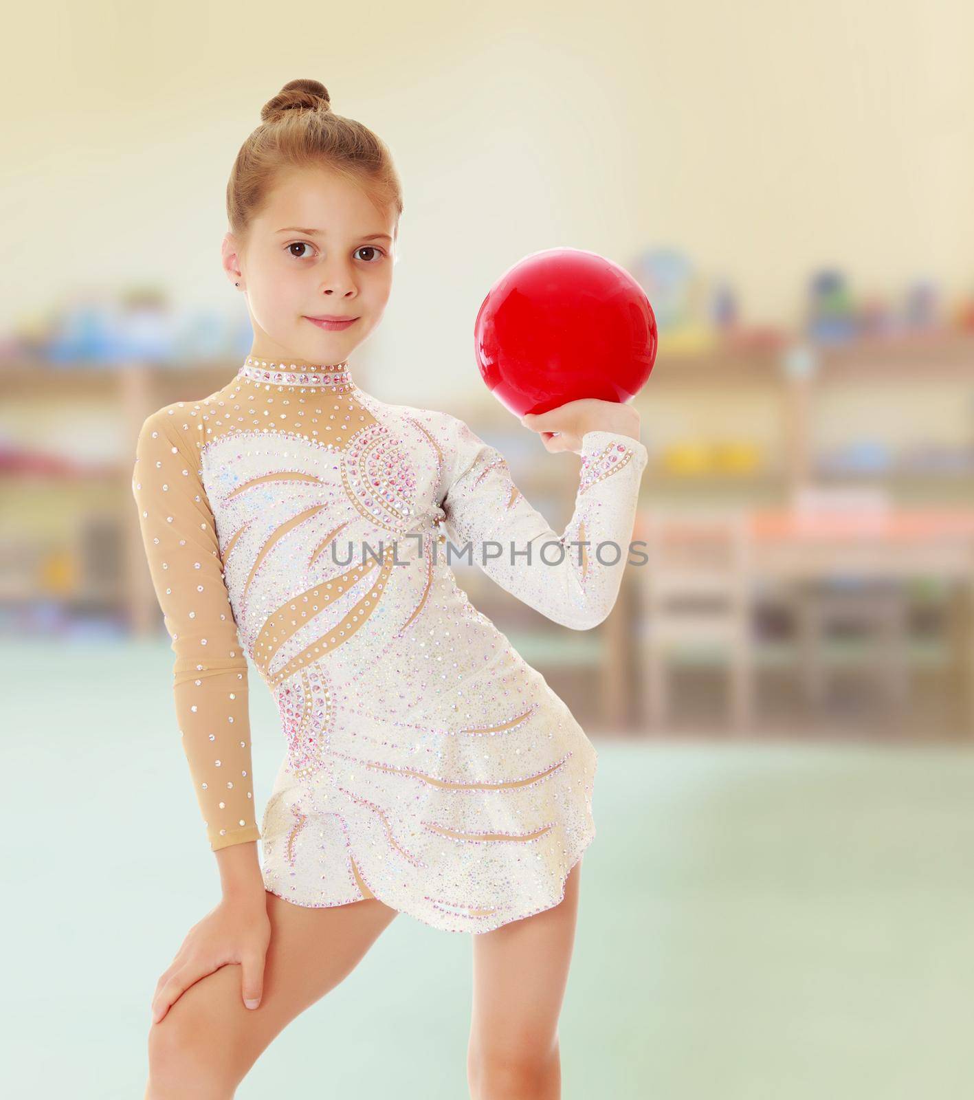 Against the background of a child's room .Beautiful little girl gymnast in elegant dress, posing with a red ball.