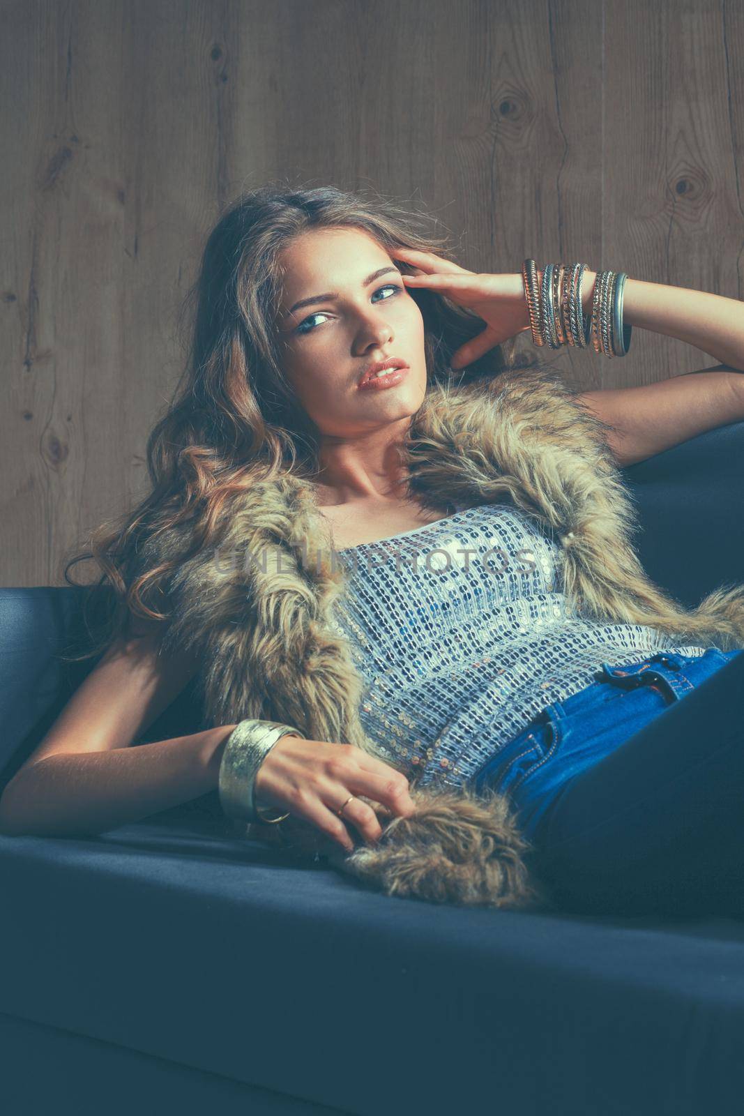 Portrait of elegant woman sitting on black sofa wearing a blue jeans and fur vest.