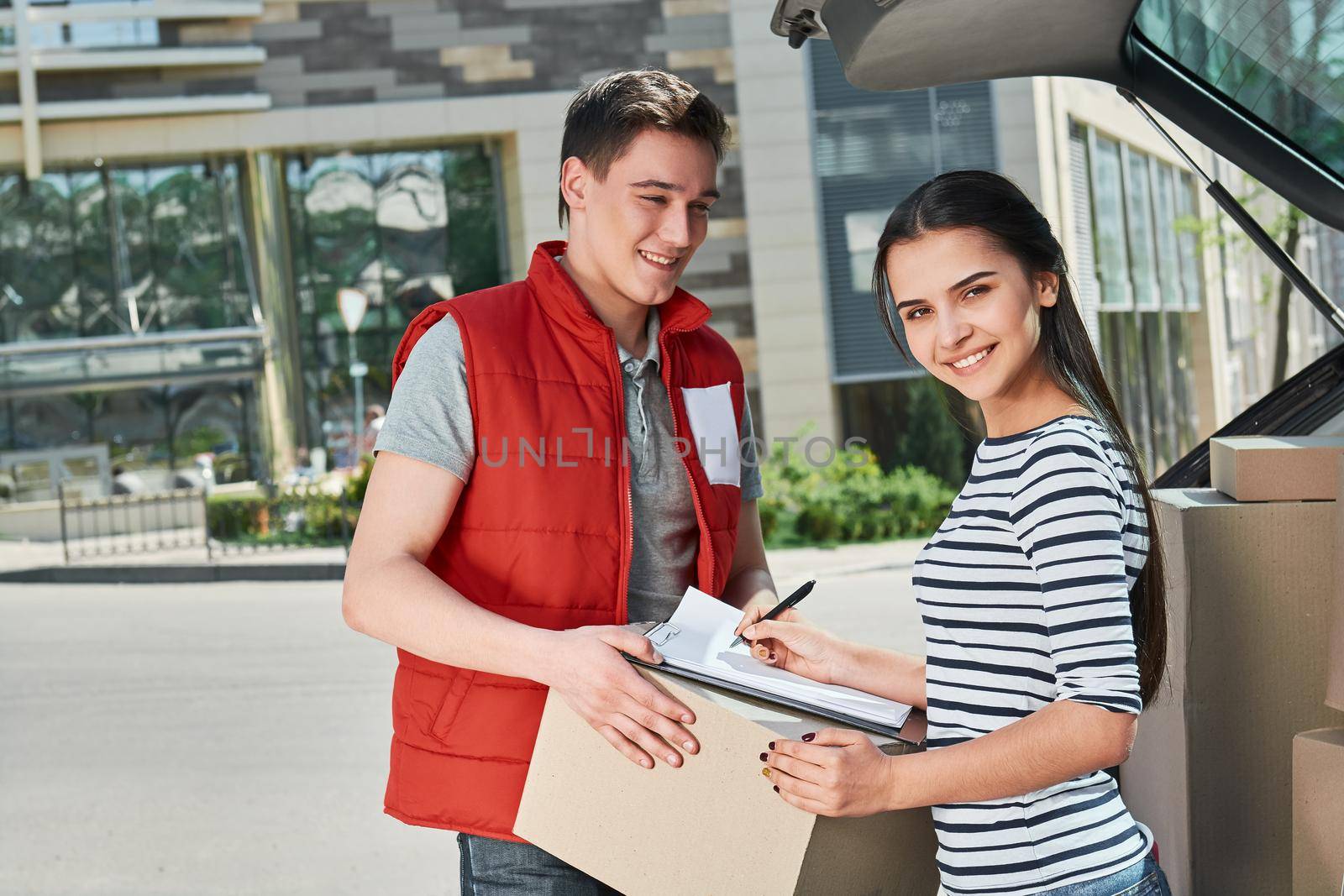 Cheerful man wearing red postal uniform is delivering parcel to a satisfied client. He brought her a parcel directly to the trunk of her car. Dark-haired woman is signing receipt of delivery package. Friendly worker, high quality delivery service. Outdoors.