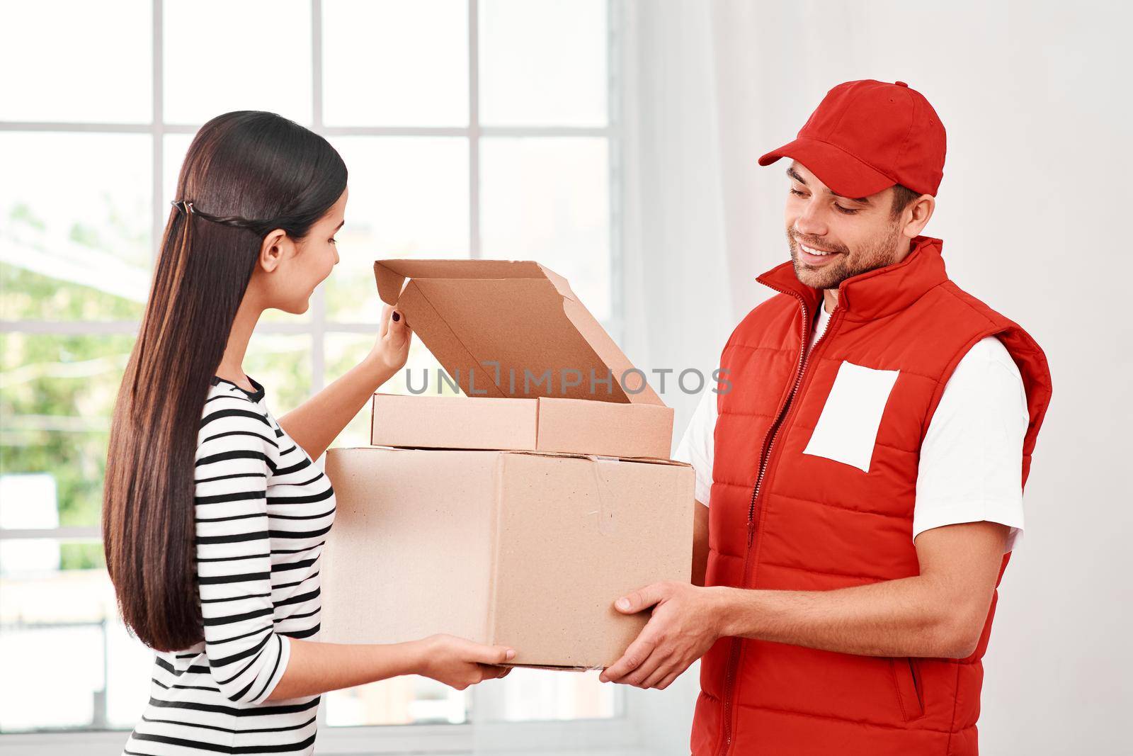Cheerful postman wearing red postal uniform is delivering parcels to a satisfied client. He brings carton boxes to its owner. Dark-haired woman opens parcel, she is glad to receive it. Friendly worker, high quality delivery service. Indoors. Bright interior.