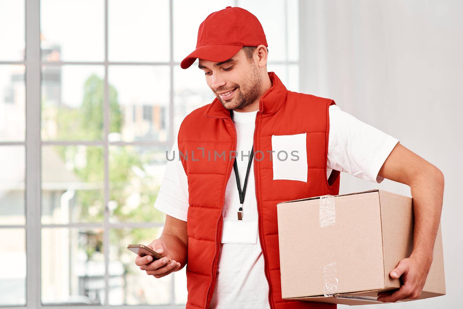 Busy postman wearing red postal uniform is delivering parcel to a client. He holds carton box and looking at his phone while searching customer number. Friendly worker, high quality delivery service. Indoors. Bright interior.