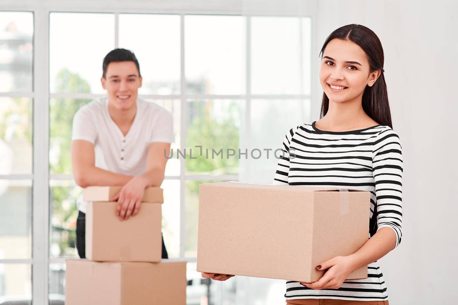 Young couple of freelancers, entrepreneurs, sellers prepare product, parcel box for delivering to customer. Woman holds carton box and looking at the camera, while man standing near stack of boxes in the background. Indoors, bright interior. Online selling, e-commerce concept