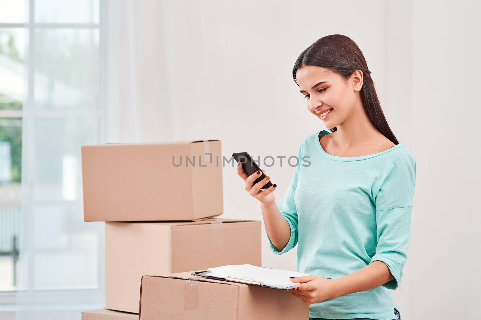 Young entrepreneur, SME, freelance woman working online, business by using smartphone, making purchase, order and preparing packages. Woman standing near stack of boxes, holding a clipboard and looking at her phone