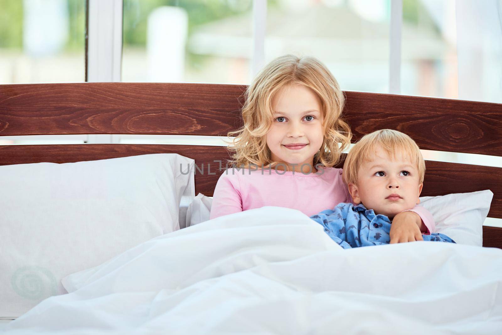 Portrait of cure little sister and brother in pajamas looking at camera and smiling while lying together in a large bed at home. Quarantine. Spending time together. Family