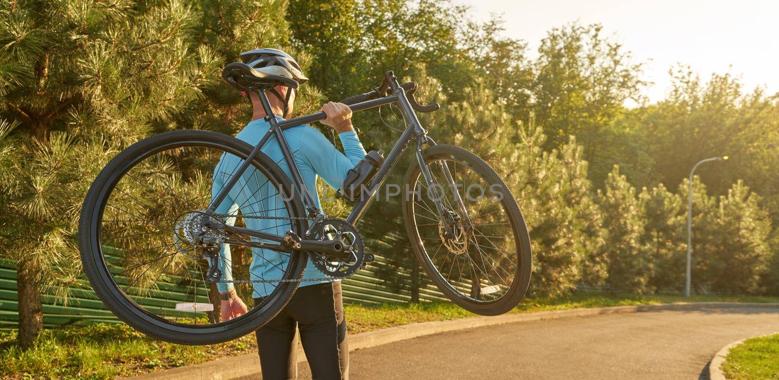 Strong athletic man in sportswear holding a bicycle while standing in park at sunset, cycling outdoors and enjoying amazing nature view. Active lifestyle and sport