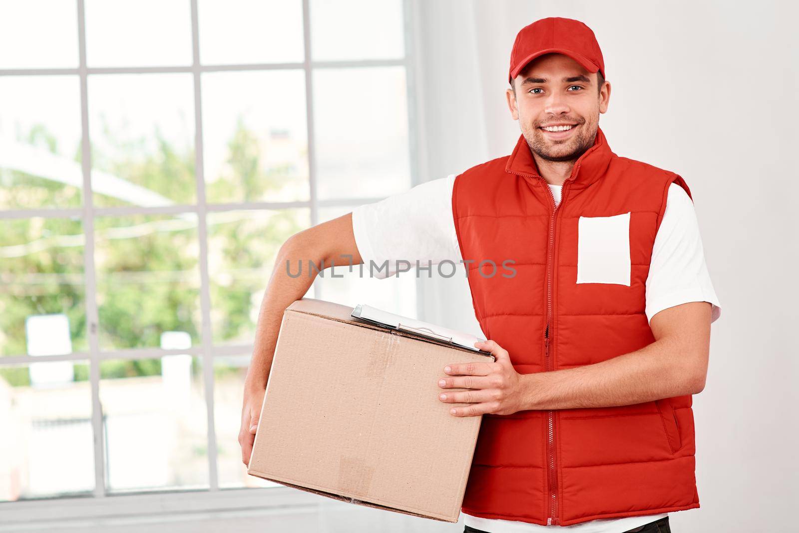 Cheerful postman wearing red postal uniform is delivering parcel to a client. He holds carton box and looking at the camera with a smile. Friendly worker, high quality delivery service. Indoors. Bright interior.