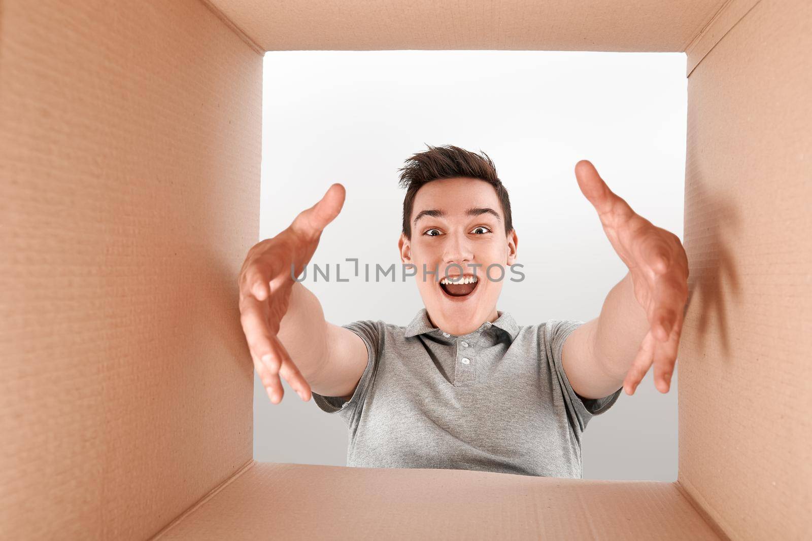 Cheerful boy wearing grey T-shirt unpacking, opening carton box. He stretches his arms forward to get a purchase out of the box. He is delighted with the speed of delivery of goods
