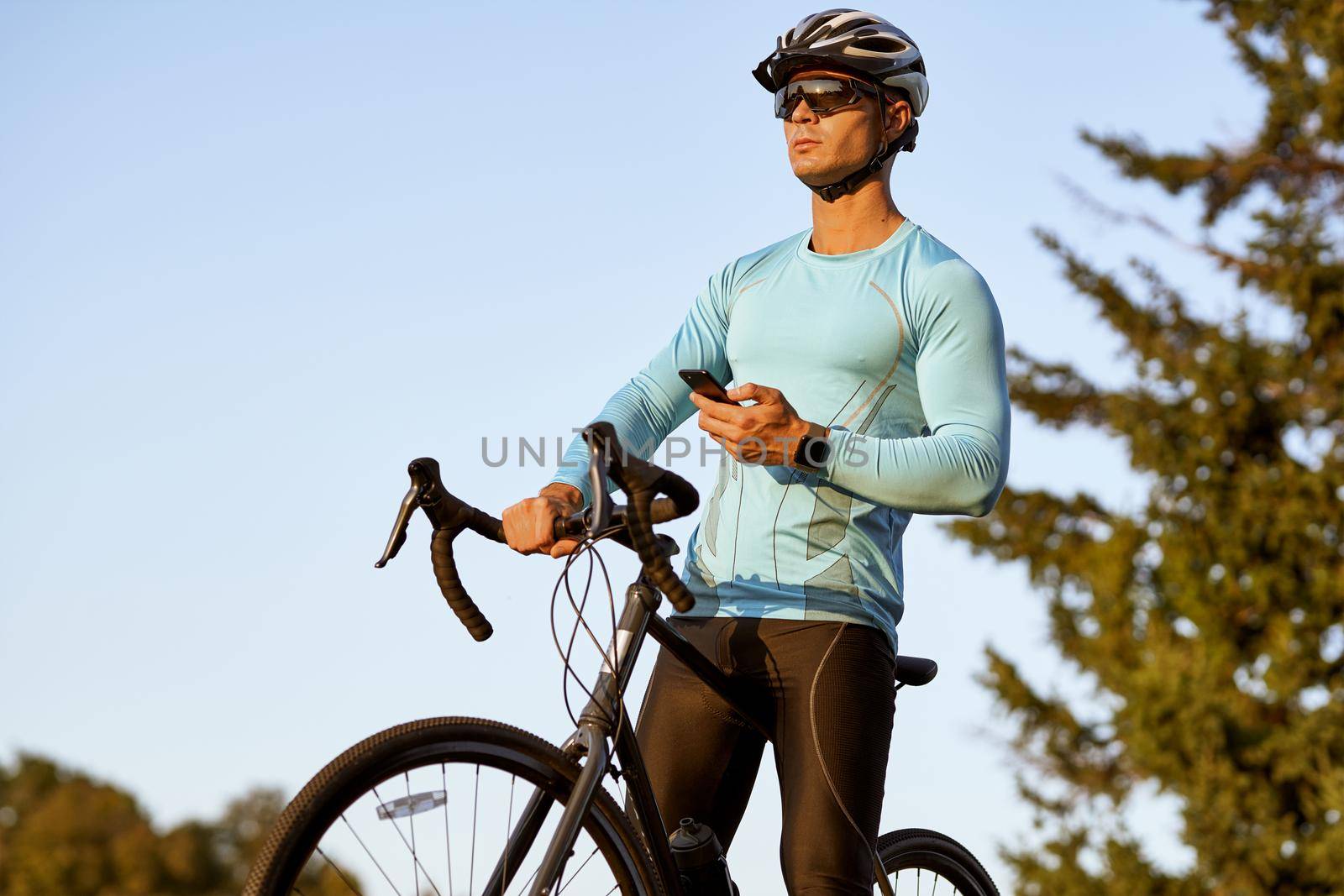 Professional male cyclist in sportswear holding smartphone and looking away, resting after ride while standing with his bike in the park on a sunny day. Active lifestyle and sport