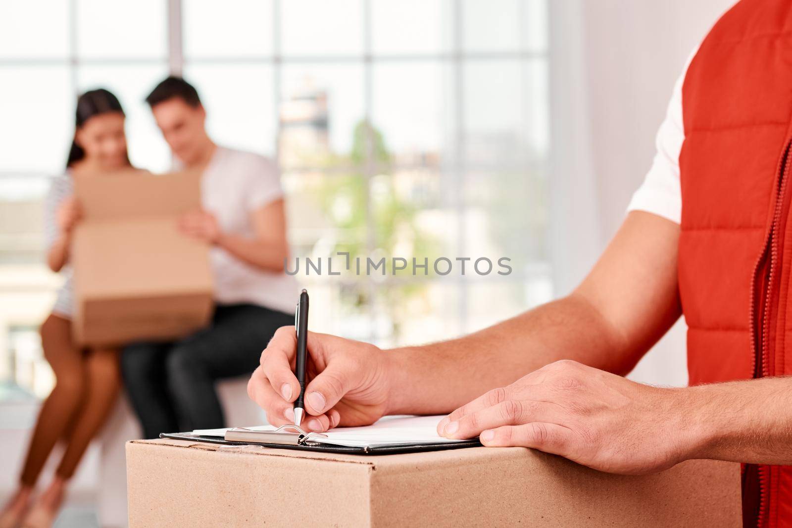 Close-up of a delivery man in red postal uniform filling in document, holding it on a carton box. Happy couple unpacking their parcel in the background. Busy worker, high quality delivery service. Indoors. Bright interior.