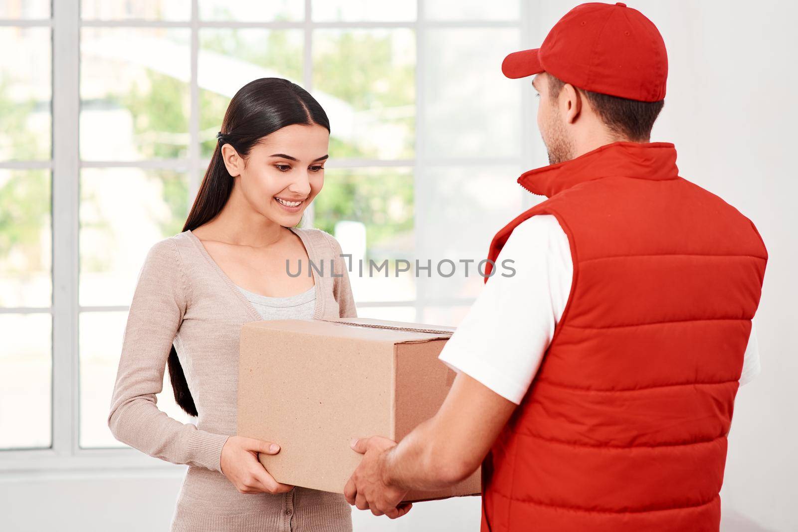 Cheerful postman wearing red postal uniform is delivering parcel to a satisfied client. He brings carton box to its owner. Dark-haired woman is taking parcel, she is glad to receive it. Friendly worker, high quality delivery service. Indoors. Bright interior.