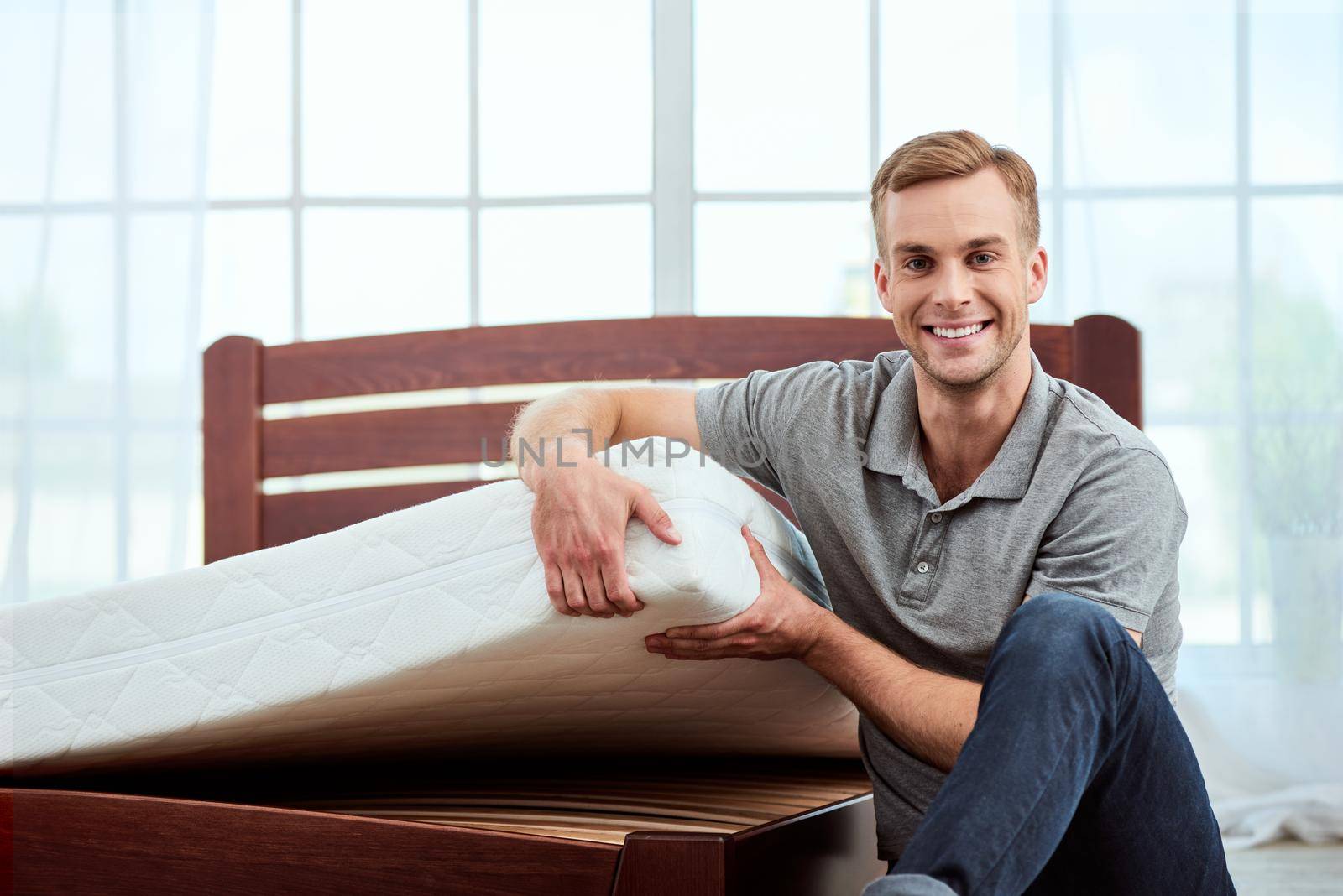 Perfect choice. Young and happy handsome man showing big and comfortable orthopedic mattress at camera and smiling by friendsstock