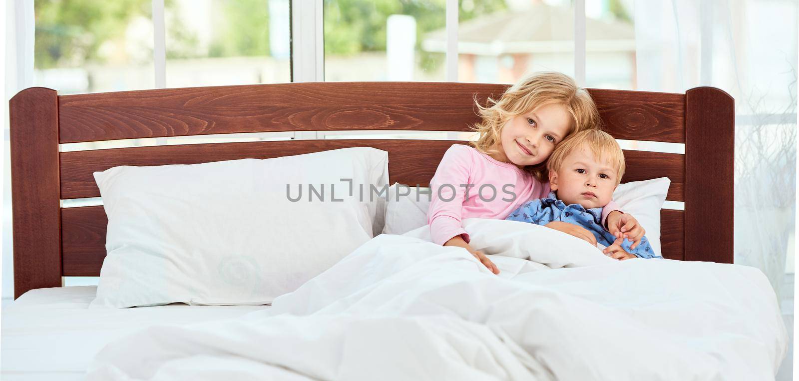 Cute little brother and sister in pajamas and looking at camera with smile while lying together in a large bed at home. Quarantine. Enjoying weekend at home. Family