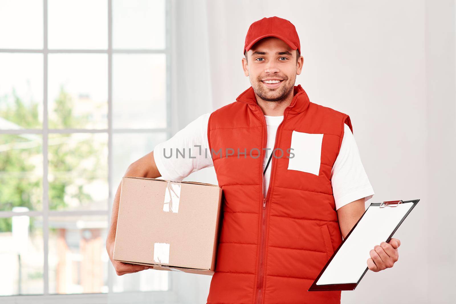 Cheerful postman wearing red postal uniform is delivering parcel to a client. He holds carton box, folder and looking at the camera with a smile. Friendly worker, high quality delivery service. Indoors. Bright interior.