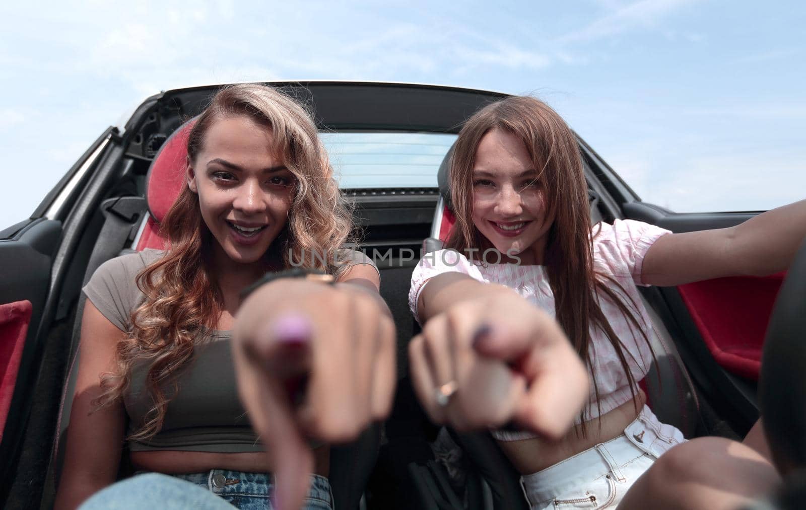 two happy girlfriends sitting in a car and pointing at you by asdf