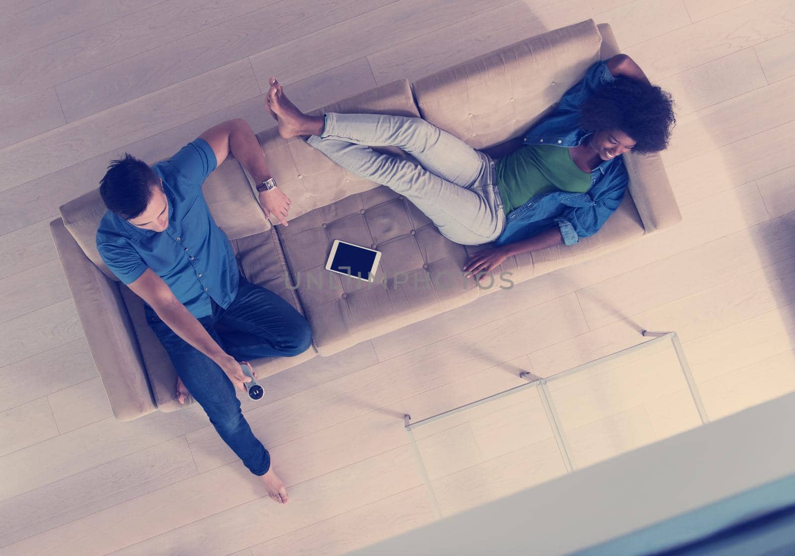 Young multiethnic couple relaxes on the sofa in the luxury living room, using a tablet and remote control top view