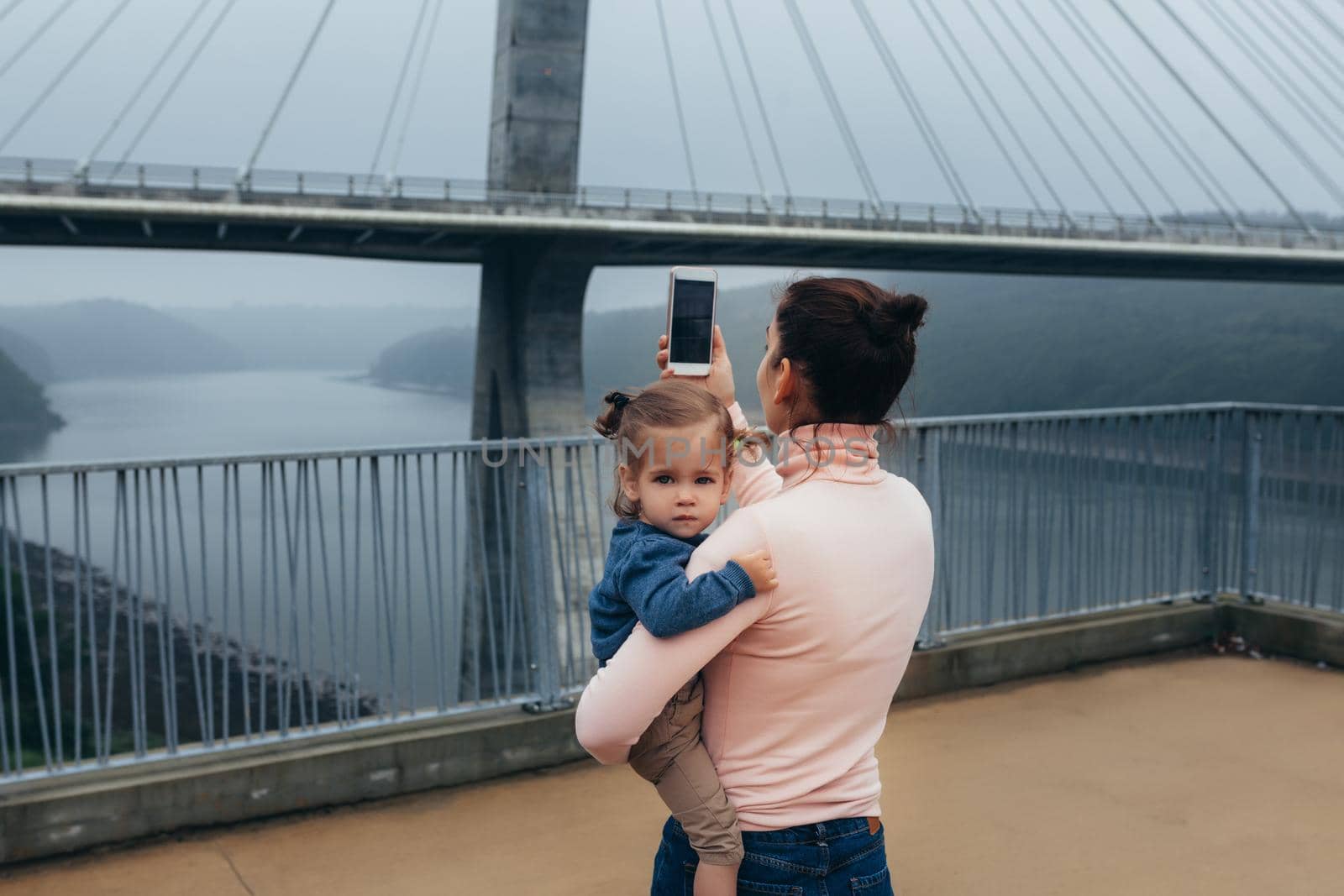 Mother and little daughter traveling together Mother photographing bridge while little girl sitting on hands France Bresta city. Caucasian family with one child standing observation deck take a photo