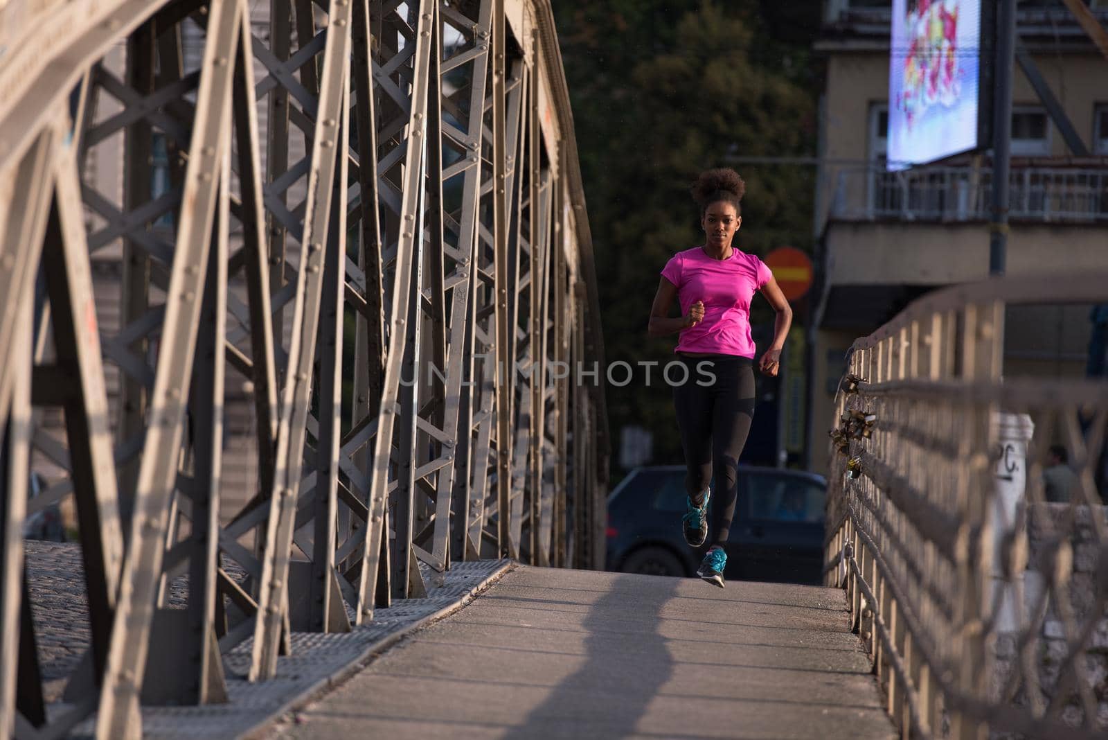 Young sporty african american woman running on sidewalk across the bridge at early morning jogging with city sunrise scene in background