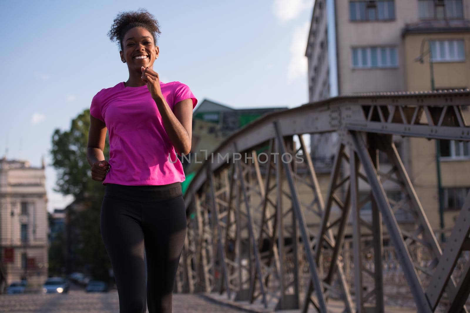 Young sporty african american woman running on sidewalk across the bridge at early morning jogging with city sunrise scene in background