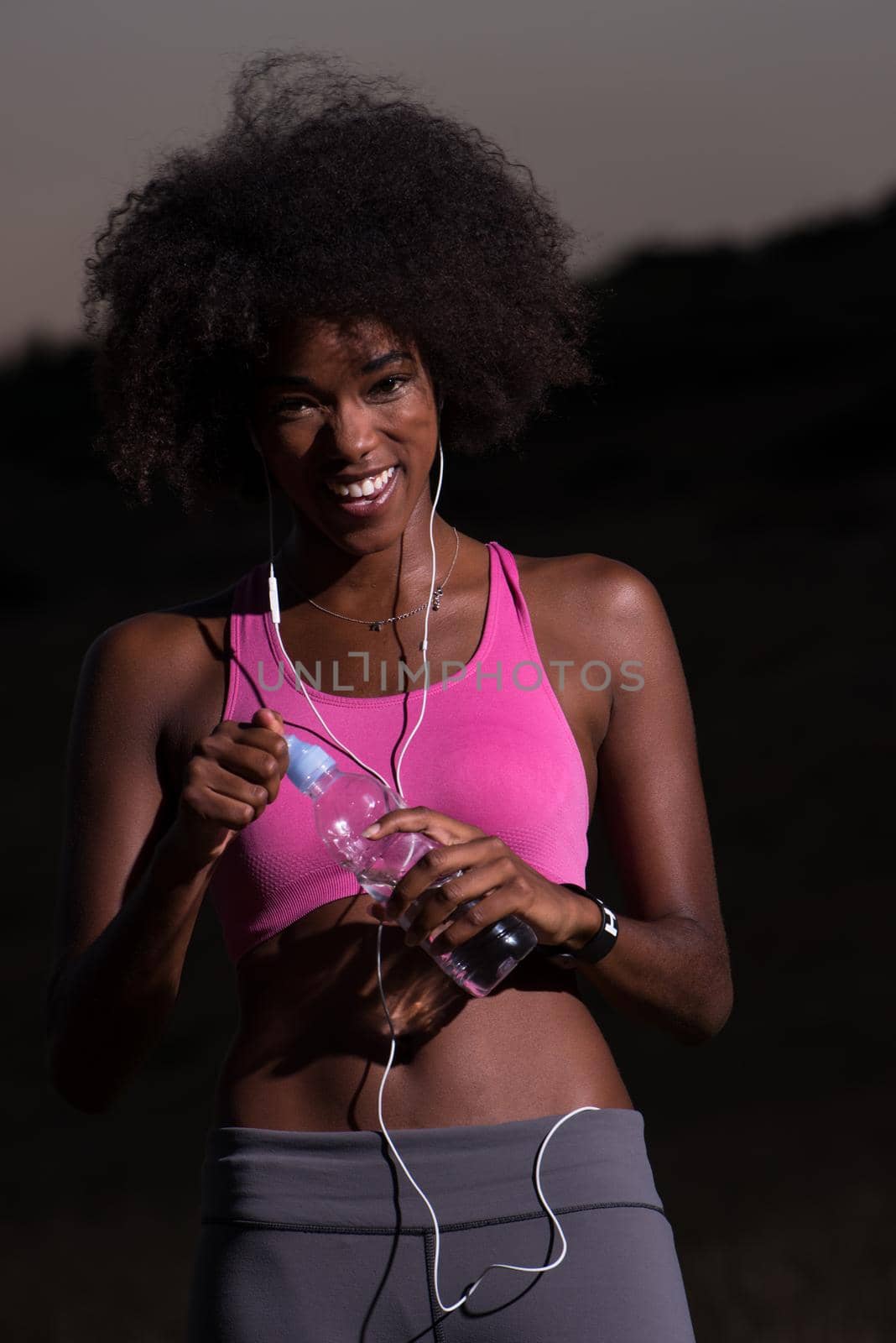 portrait of a young beautiful african american woman with headphones and a bottle of water after jogging in nature beautiful summer night