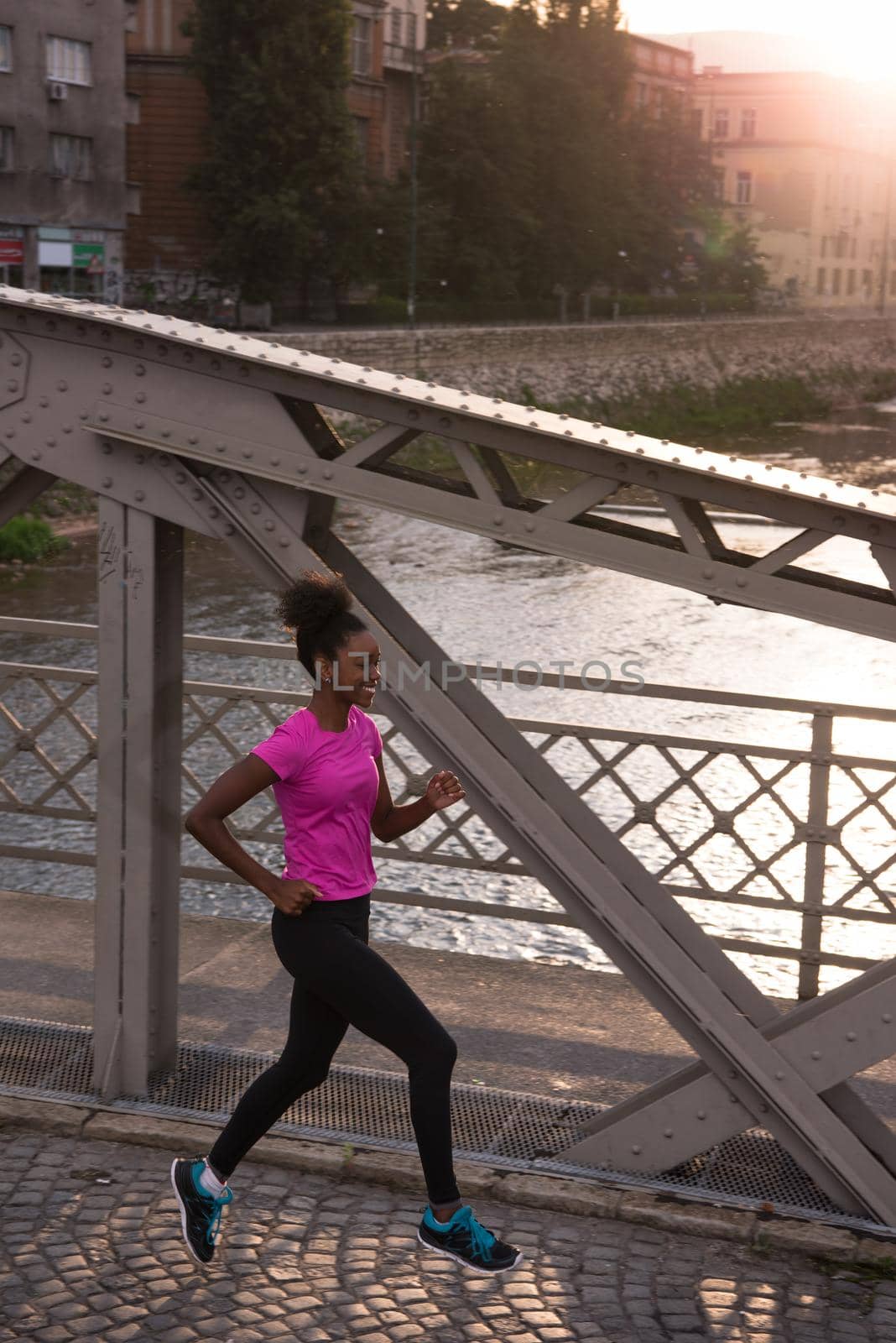 Young sporty african american woman running on sidewalk across the bridge at early morning jogging with city sunrise scene in background