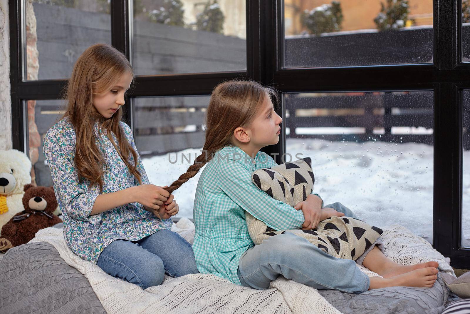 Two beautiful caucasian sisters sitting on the windowsill, they look out of the window. Older sister cares for younger sister's hair