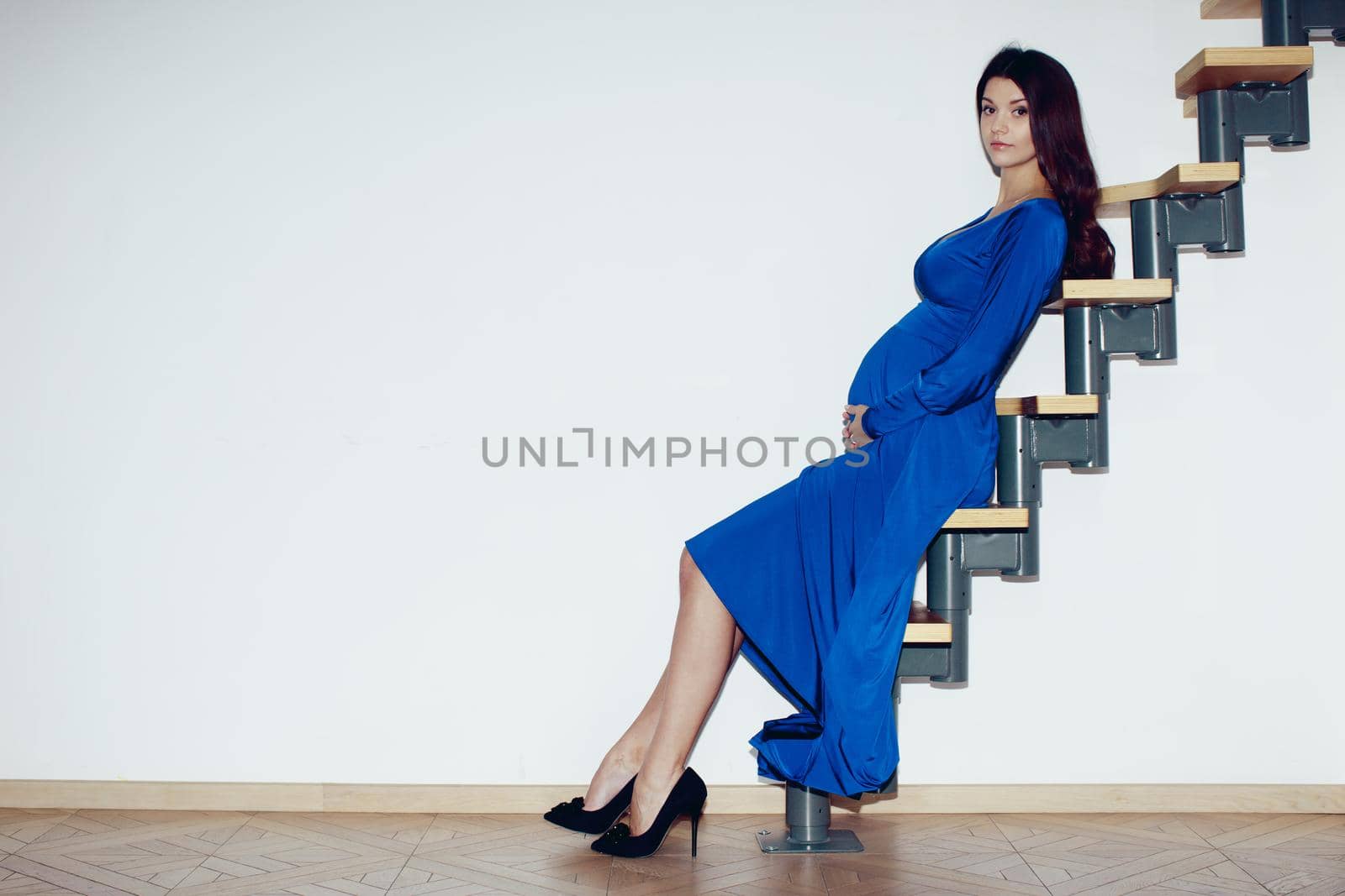 Beautiful pregnant girl, brunette, big breasts, hands on belly, blue dress, sitting on a ladder against a white wall in the interior
