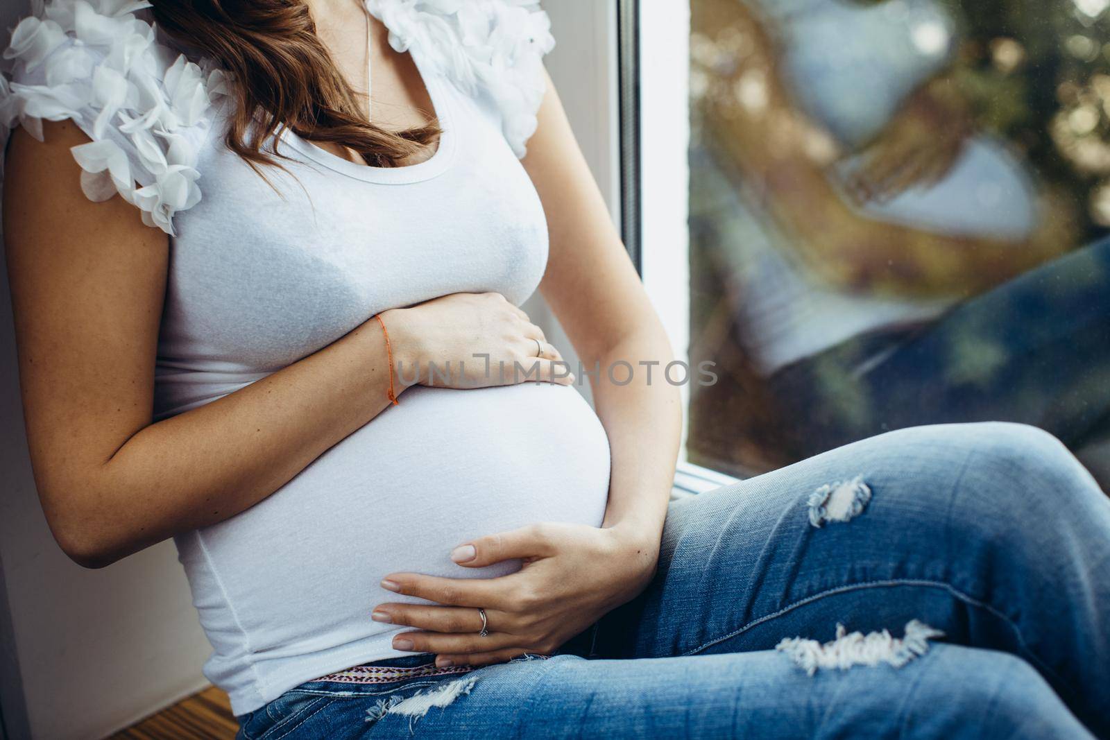 Belly closeup of a future mother. Pregnant brunette girl sitting on the windowsill, holding his stomach outside, the greens, the girl white T-shirt and ripped jeans.