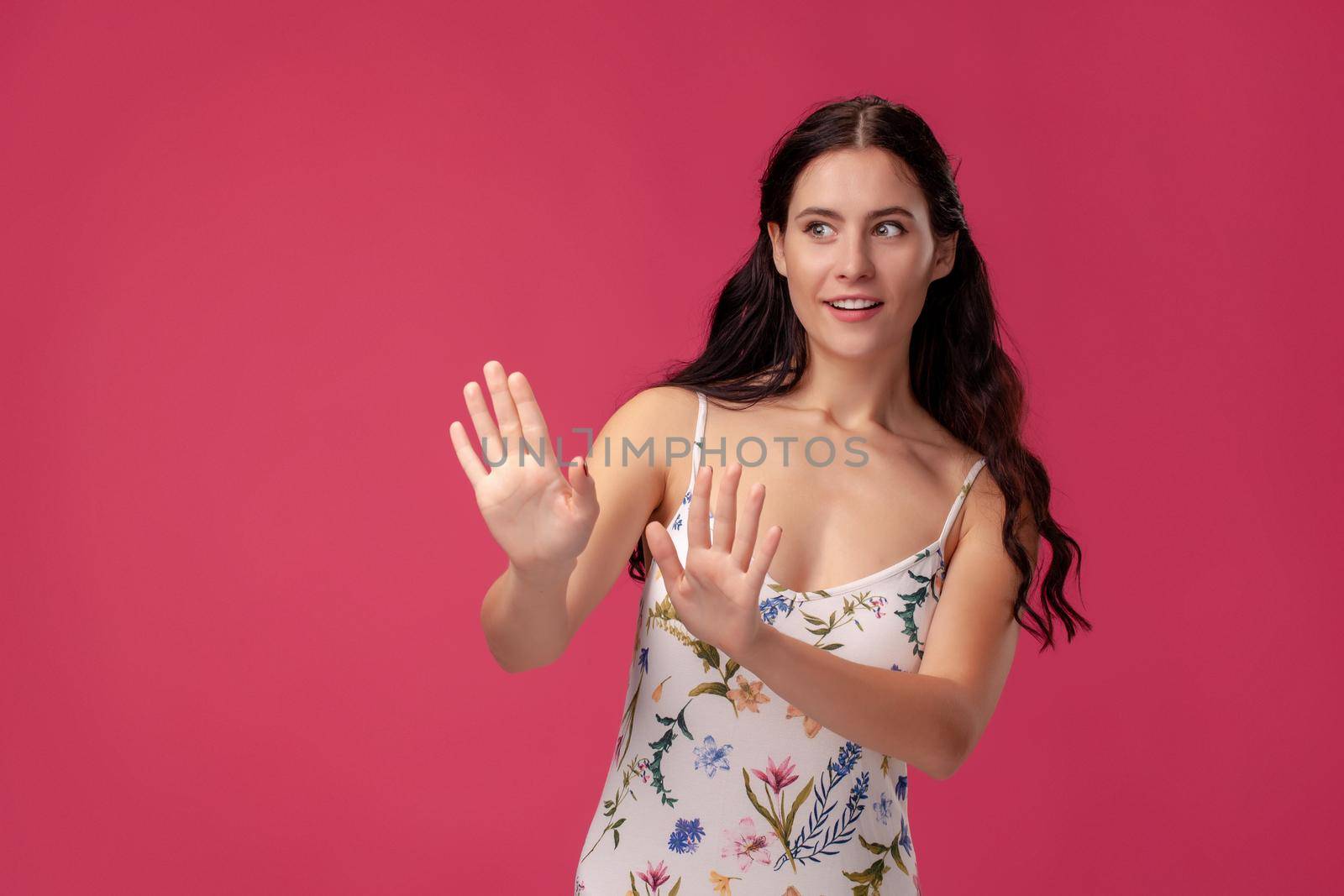 Portrait of a young pretty female in a white dress with floral print standing on a pink wall background in studio. She is acting like saying no to someone. People sincere emotions, lifestyle concept. Mockup copy space.