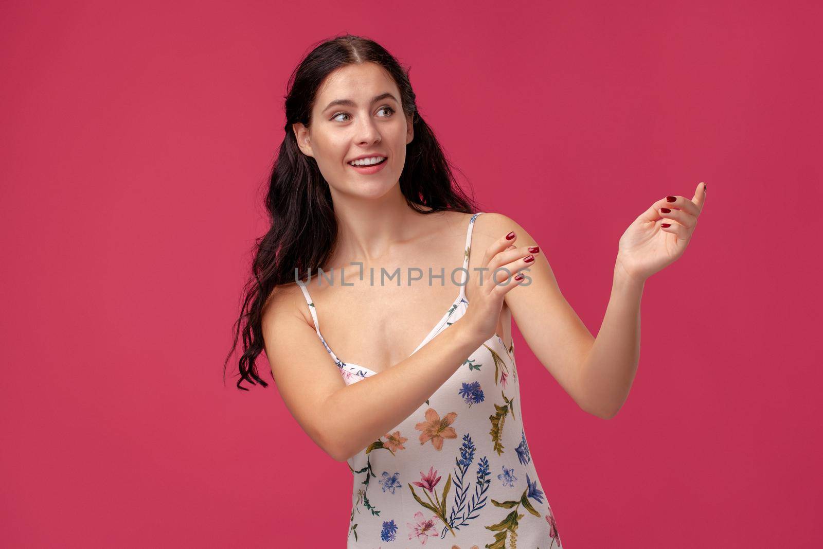 Portrait of a beautiful lady in a white dress with floral print standing on a pink wall background in studio. She is pointing at something, smiling and looking happy. People sincere emotions, lifestyle concept. Mockup copy space.