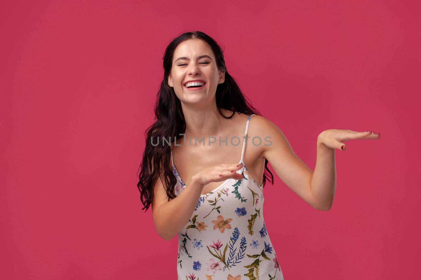 Portrait of a pretty woman in a white dress with floral print standing on a pink wall background in studio. She is laughing and looking happy. People sincere emotions, lifestyle concept. Mockup copy space.