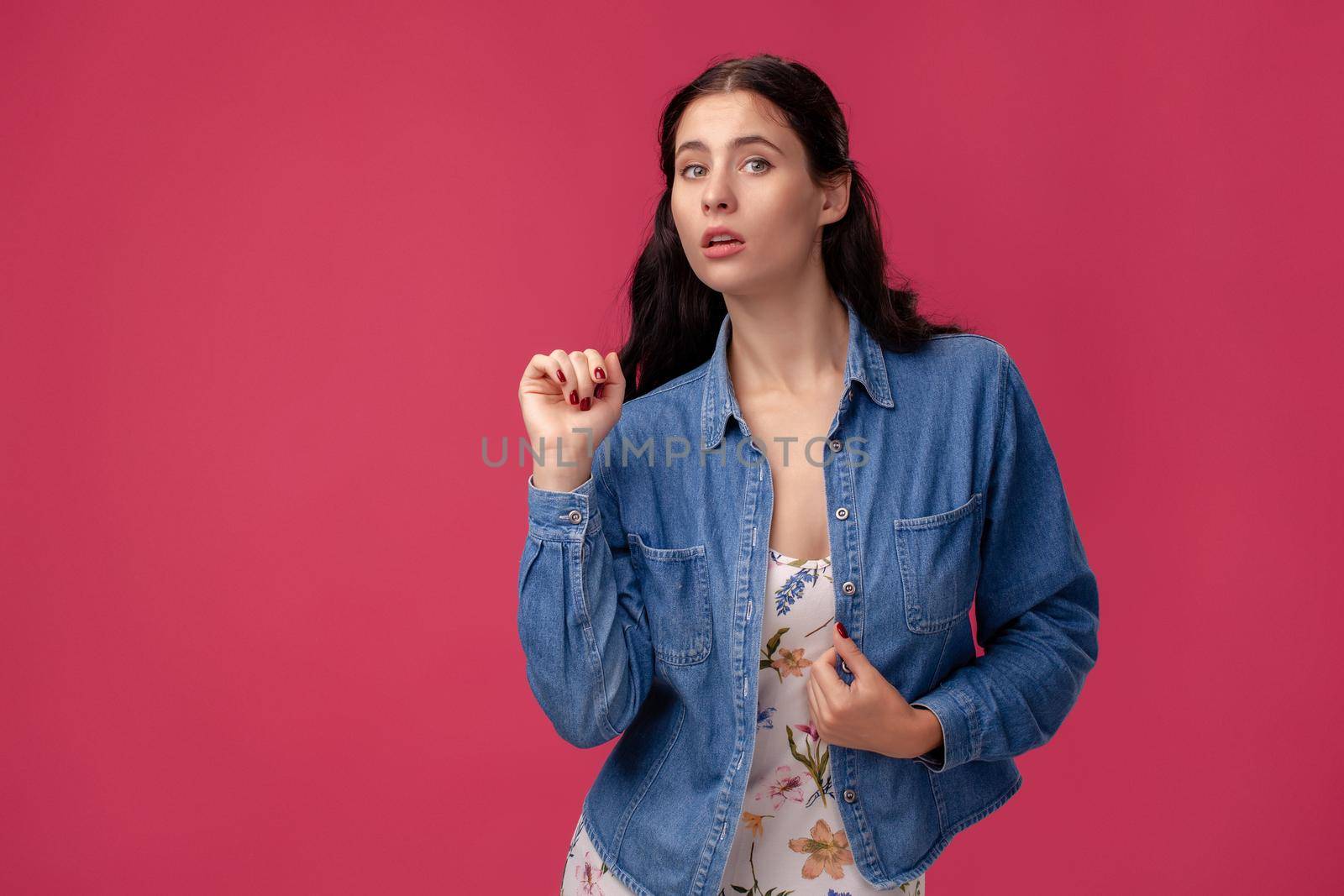 Portrait of a wondered young woman in a white dress with floral print and blue denim shirt standing on a pink wall background in studio. People sincere emotions, lifestyle concept. Mockup copy space.
