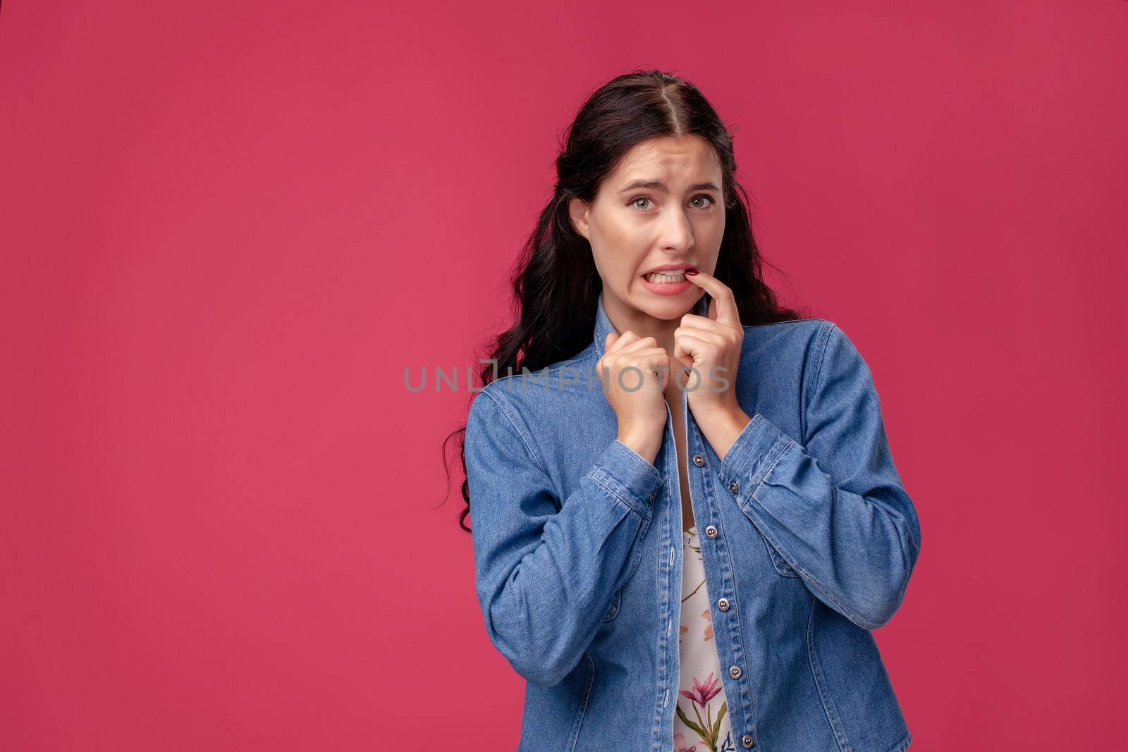 Portrait of a charming woman in a white dress with floral print. She is holding a collar of her blue denim shirt and touching her teeth standing on a pink wall background in studio. People sincere emotions, lifestyle concept. Mockup copy space.